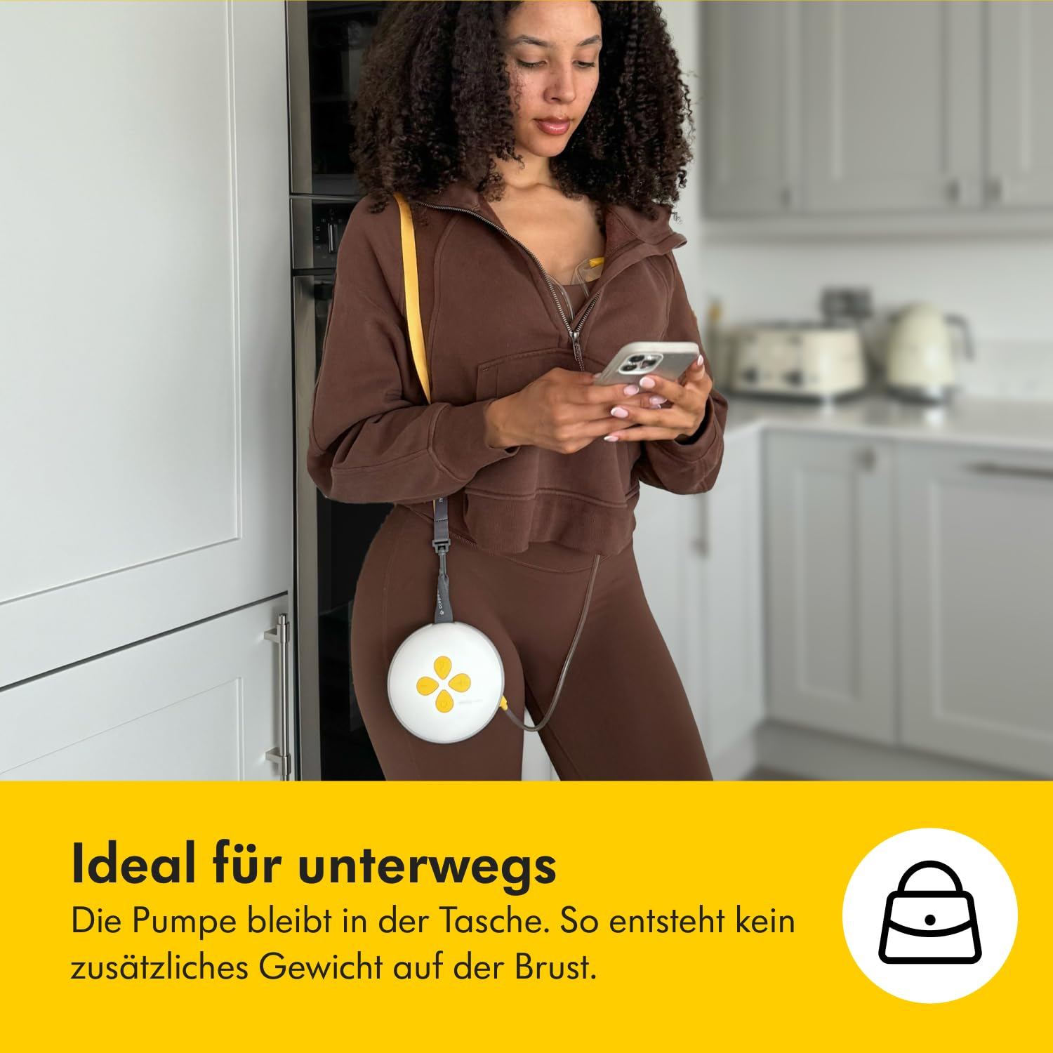 Frau in braunem Outfit hält ein Telefon und trägt eine Brustpumpe. Küche im Hintergrund. Die Pumpe bleibt in der Tasche, kein zusätzliches Gewicht.