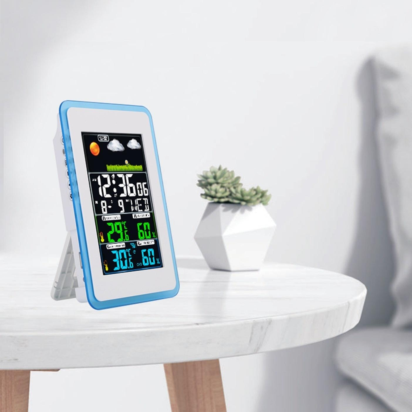 Eine digitale Wetterstation zeigt Zeit, Temperatur und Luftfeuchtigkeit an. Sie steht auf einem weißen Tisch mit einer kleinen Pflanze, neben einem weißen Sofa.