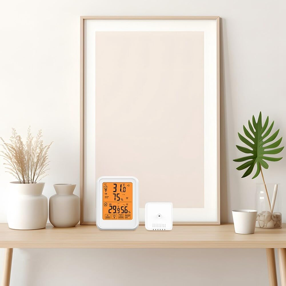 Eine weiße Wand mit einem gerahmten Bild, ein Tisch mit einem digitalen Thermometer und einem kleinen Gerät, Vasen und eine Pflanze.
