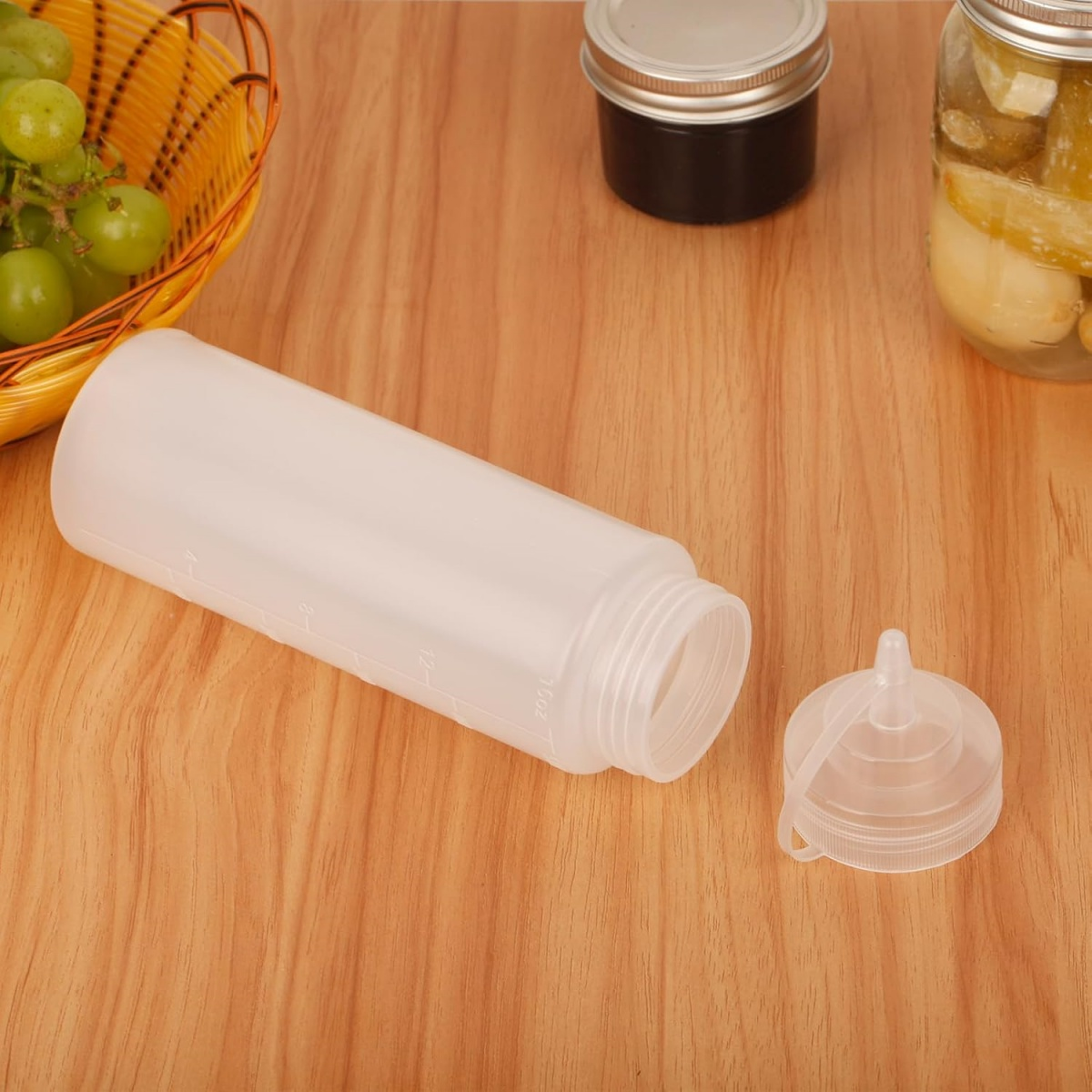 Eine weiße Quetschflasche ohne Kappe, Trauben in einem Korb und Gläser auf einer Holzoberfläche.