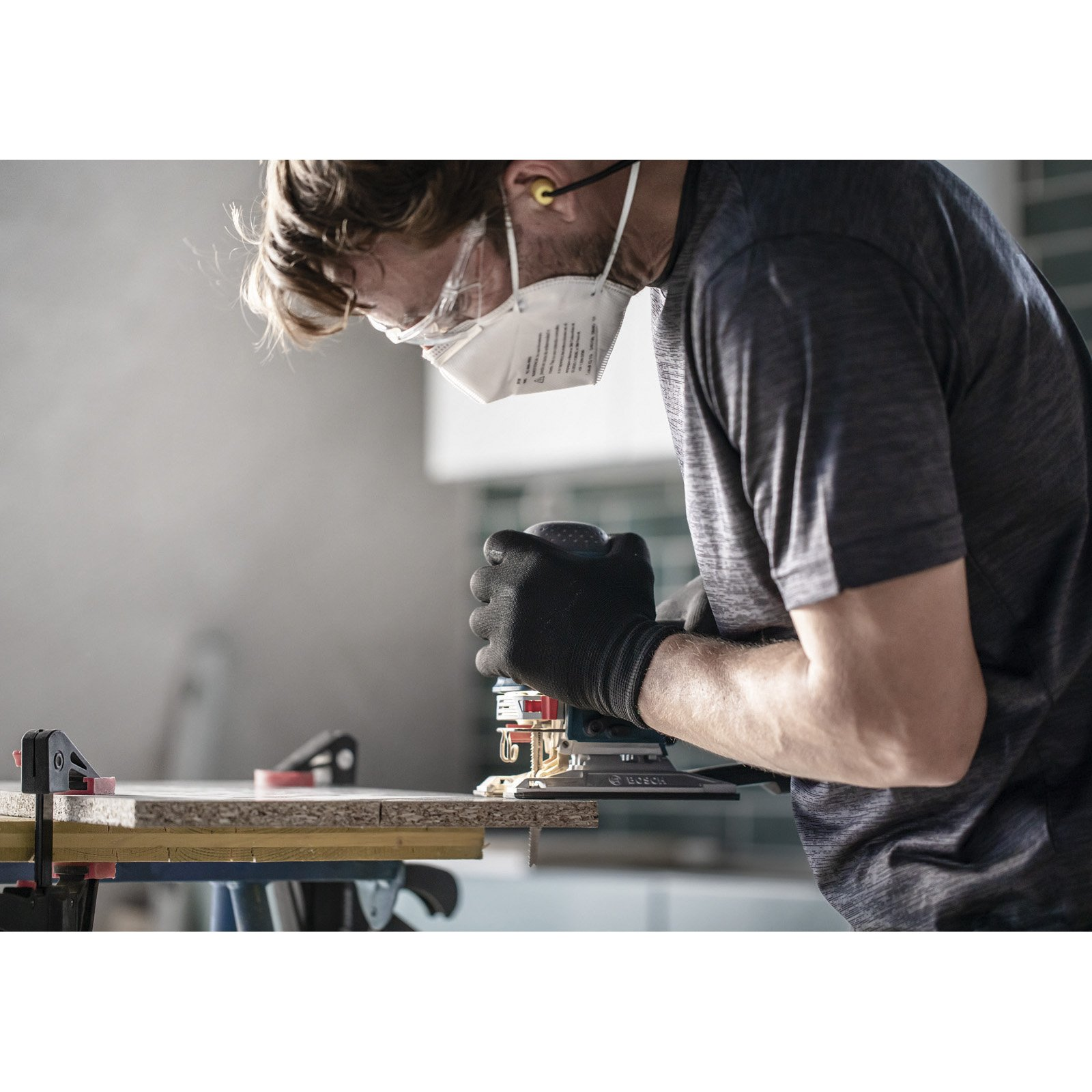 Mężczyzna szlifuje drewno szlifierką elektryczną, w rękawicach, masce i ochronie słuchu. Zacisk mocuje drewno.