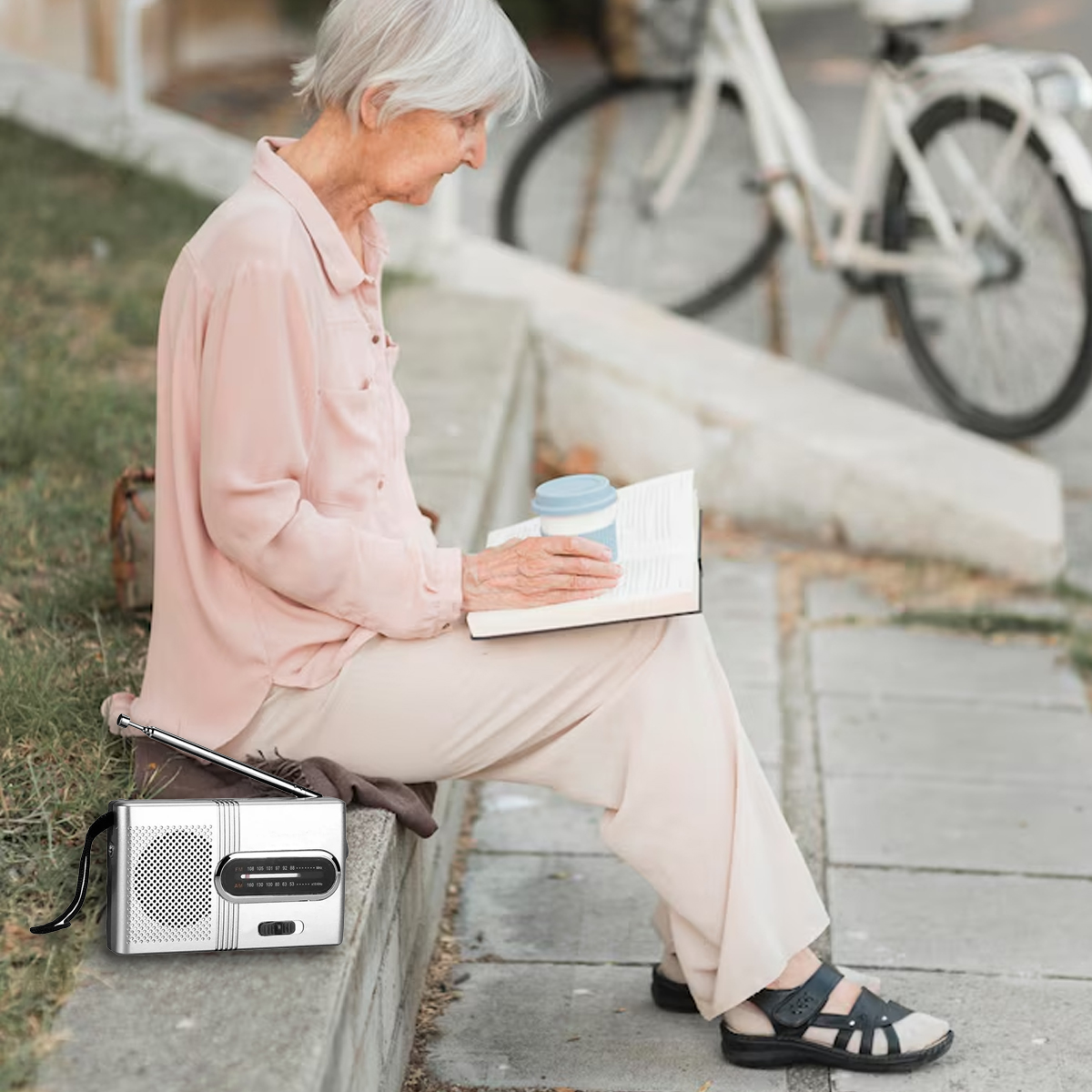 Eine ältere Frau sitzt und liest ein Buch mit einer Kaffeetasse. Ein Radio steht neben ihr. Im Hintergrund ist ein Fahrrad zu sehen.
