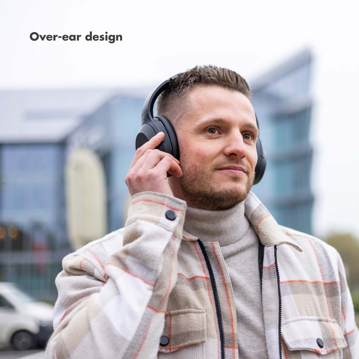 Ein Mann mit schwarzen Over-Ear-Kopfhörern. Er ist draußen, trägt ein kariertes Hemd und blickt nach oben. Moderne Gebäude sind im Hintergrund.