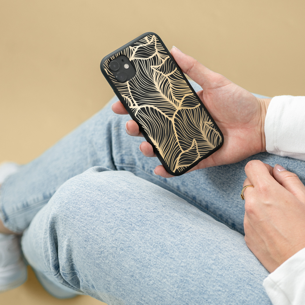 Una persona tiene in mano un telefono con una custodia nera e un design a foglie d'oro. Indossa jeans e una camicia bianca. È visibile un anello d'oro.