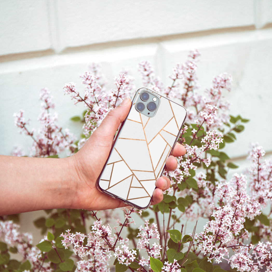 Una mano che tiene un telefono con un design geometrico bianco e oro. Fiori con fiori viola circondano la mano. Muro bianco sullo sfondo.
