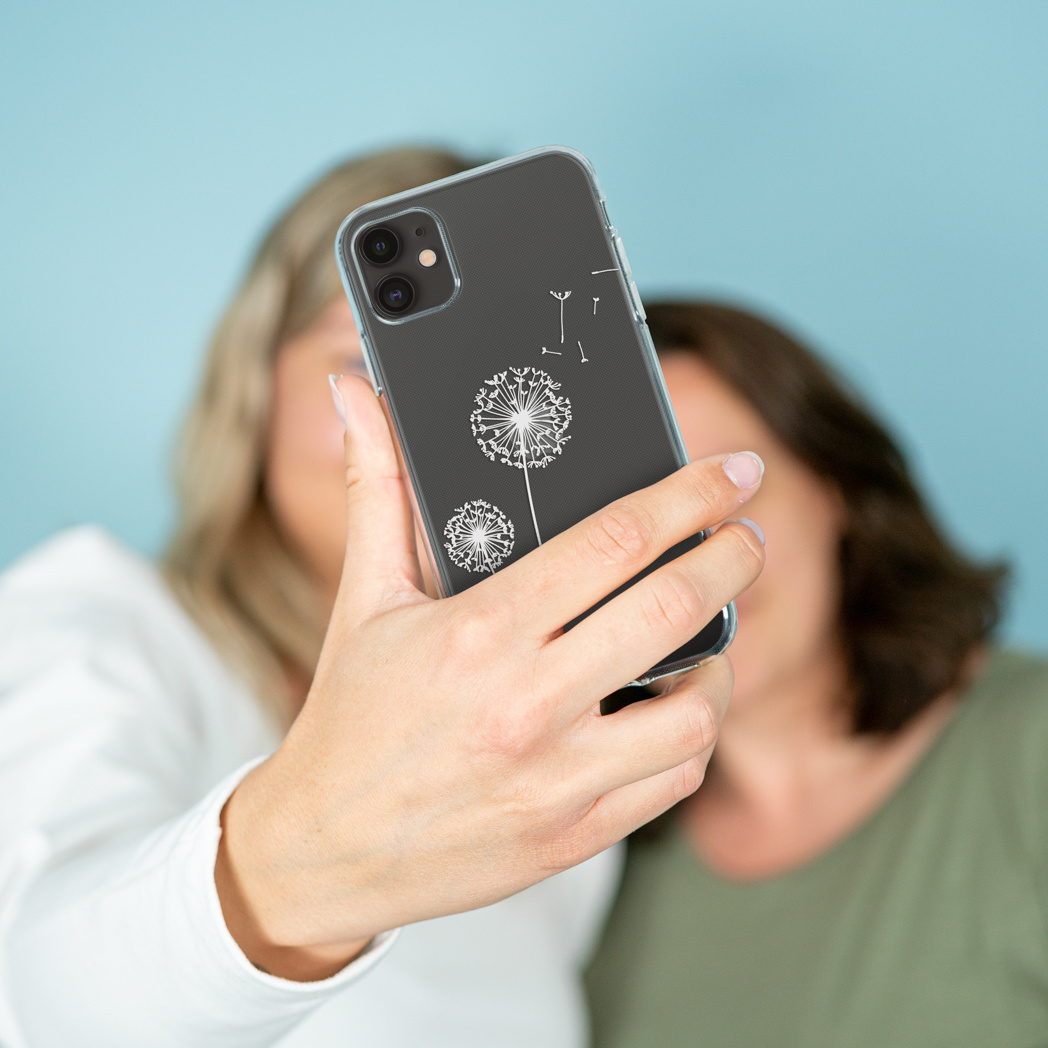 Una persona tiene in mano un telefono con un disegno di dente di leone. Due donne sfocate sullo sfondo. Il telefono è nero con una custodia trasparente.
