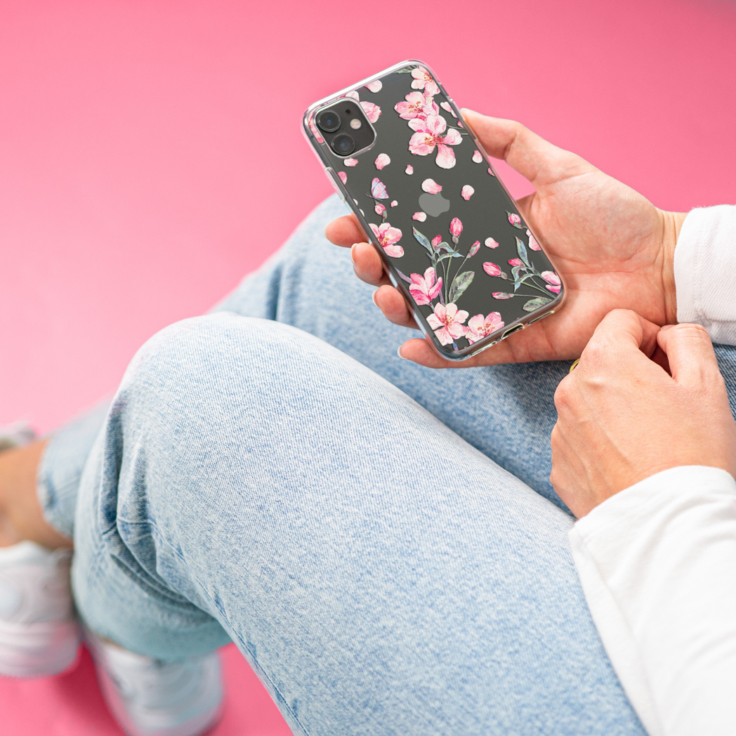 Una persona tiene in mano un telefono con una custodia floreale. Lo sfondo è rosa. La persona indossa jeans e scarpe bianche.