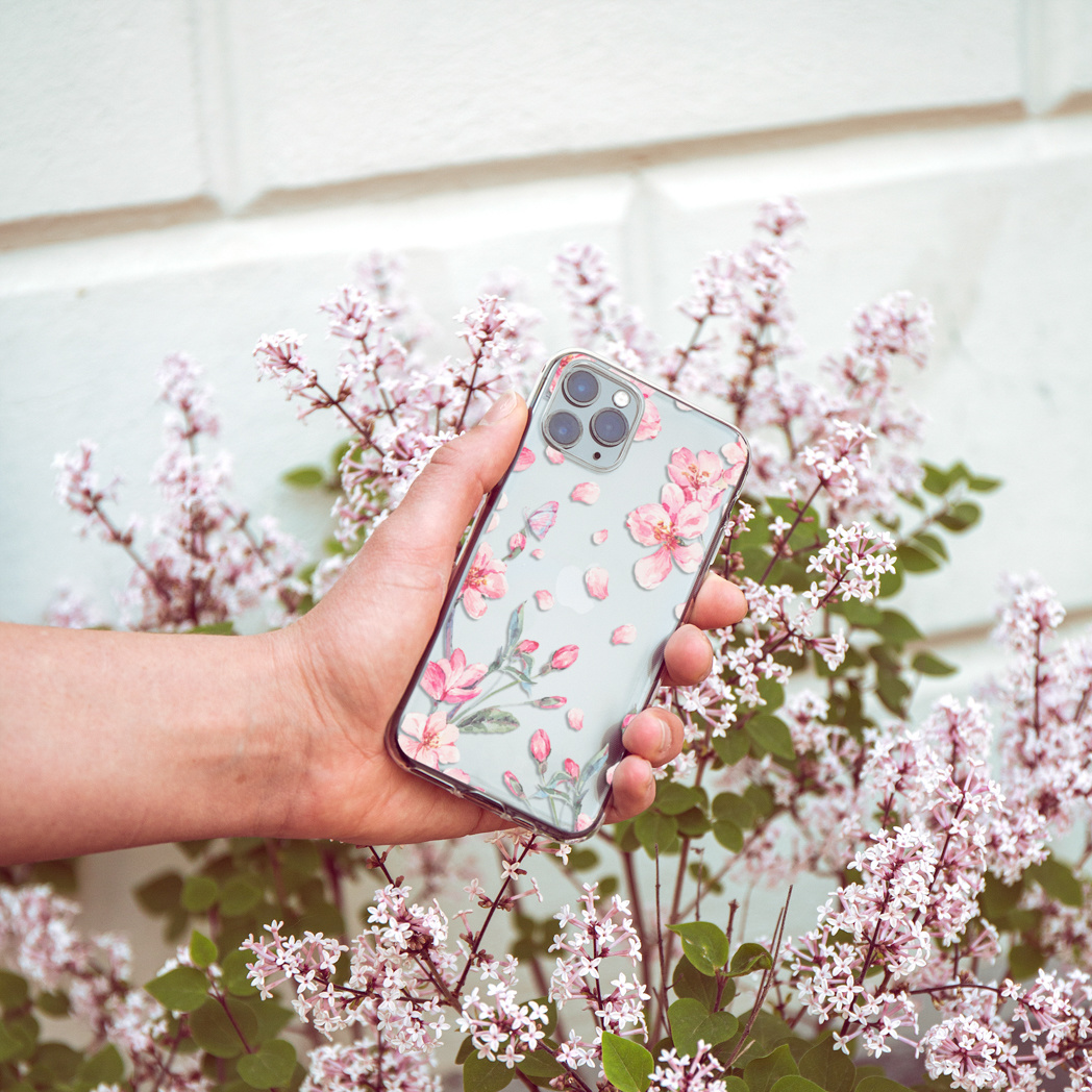 Una mano tiene in mano un telefono con custodia floreale. Fiori rosa e foglie verdi sullo sfondo. Il muro è bianco.