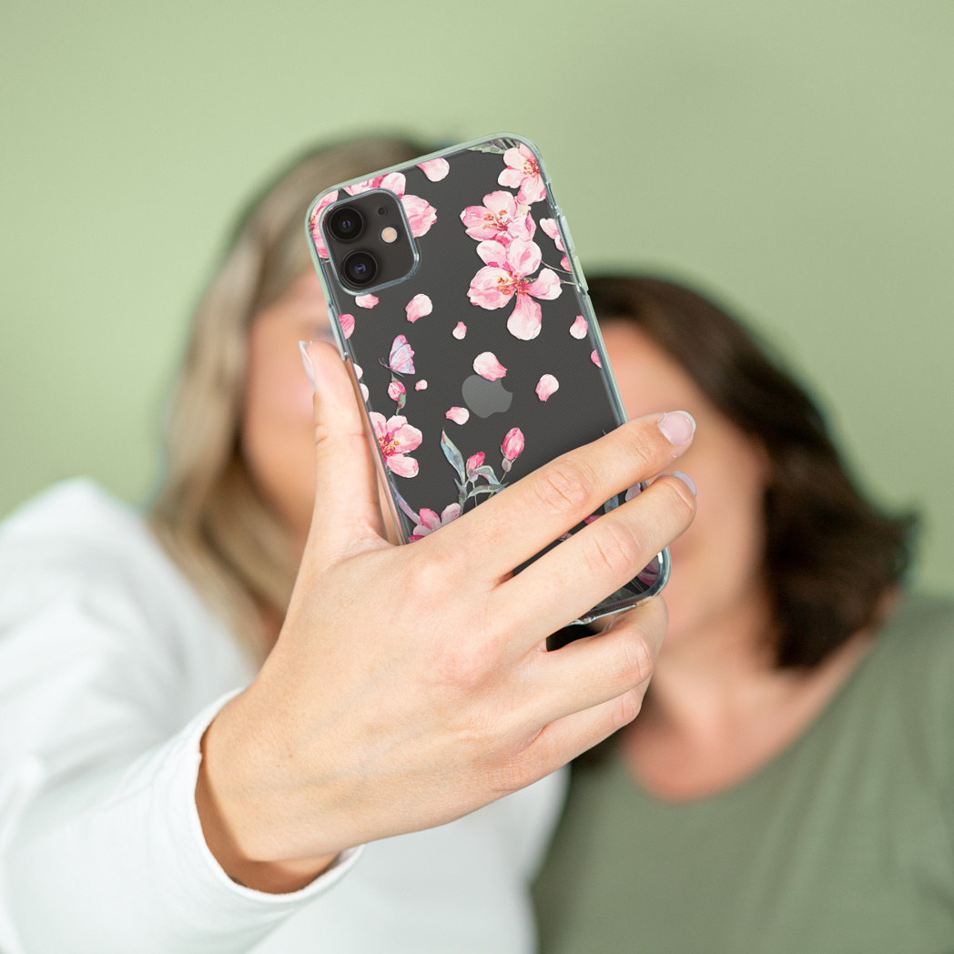 Una donna tiene in mano un telefono che mostra un motivo floreale. La custodia del telefono ha fiori rosa. Due donne sono sfocate sullo sfondo.
