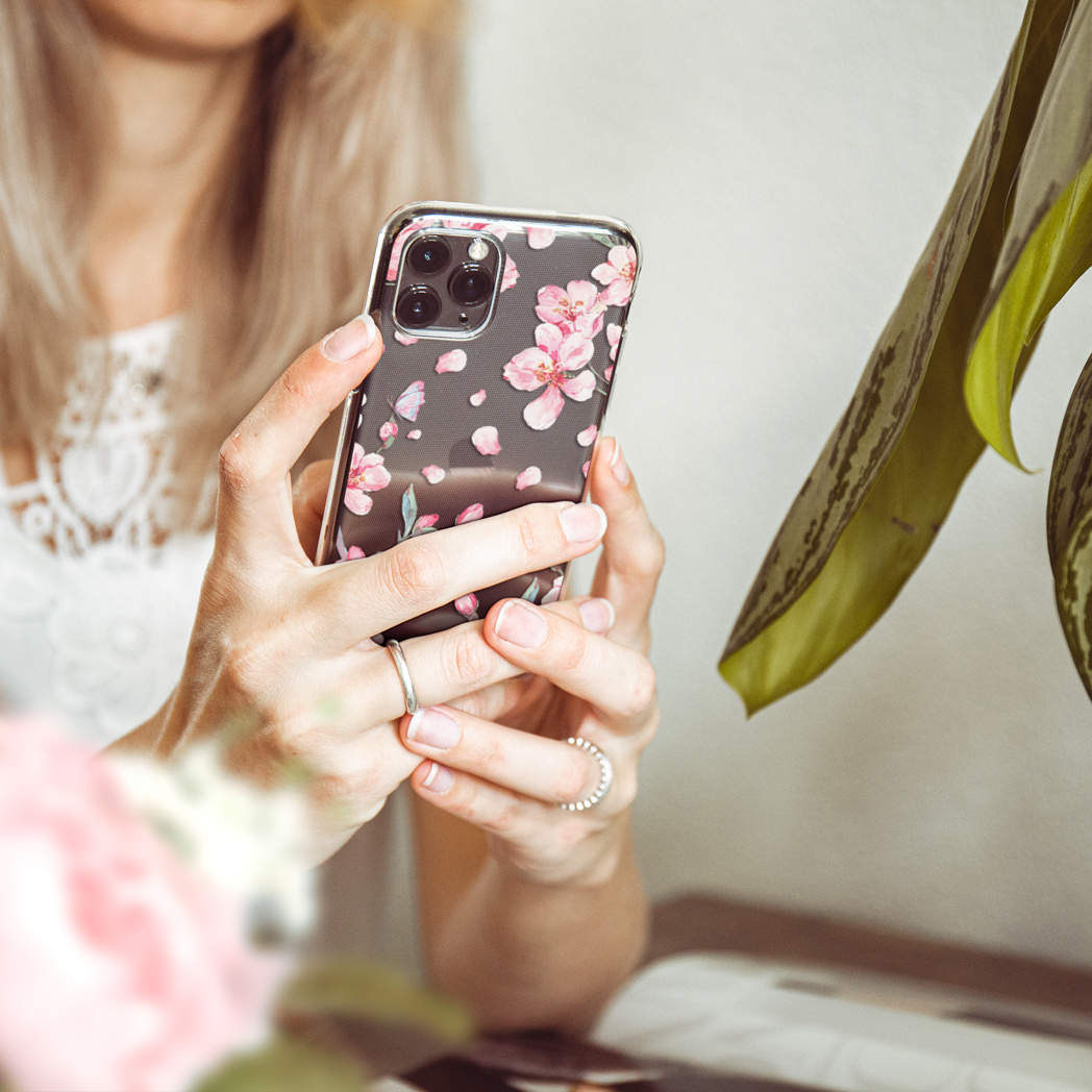 Una donna tiene in mano un telefono con una custodia floreale. Fiori rosa sulla custodia, indossa anelli. Foglie verdi sullo sfondo.