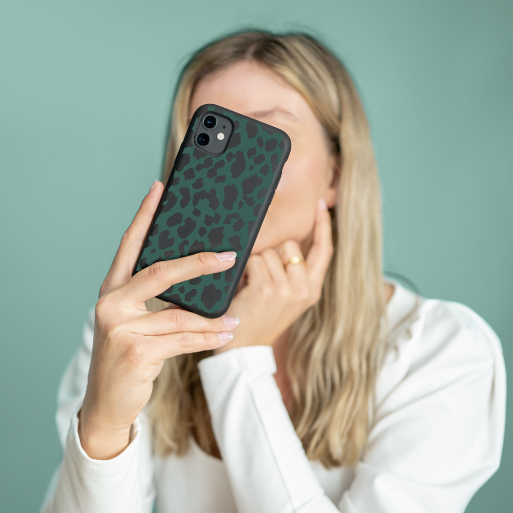 Donna bionda che tiene in mano un telefono con una custodia verde con stampa leopardata. Indossa una camicia bianca e lo sfondo è turchese.