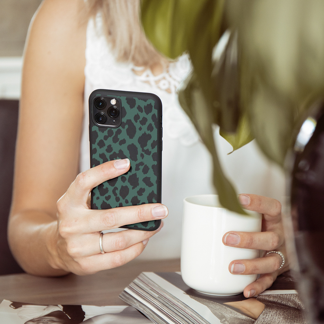 Donna che tiene un telefono con una custodia con stampa leopardata verde e una tazza bianca. Una pianta sullo sfondo.