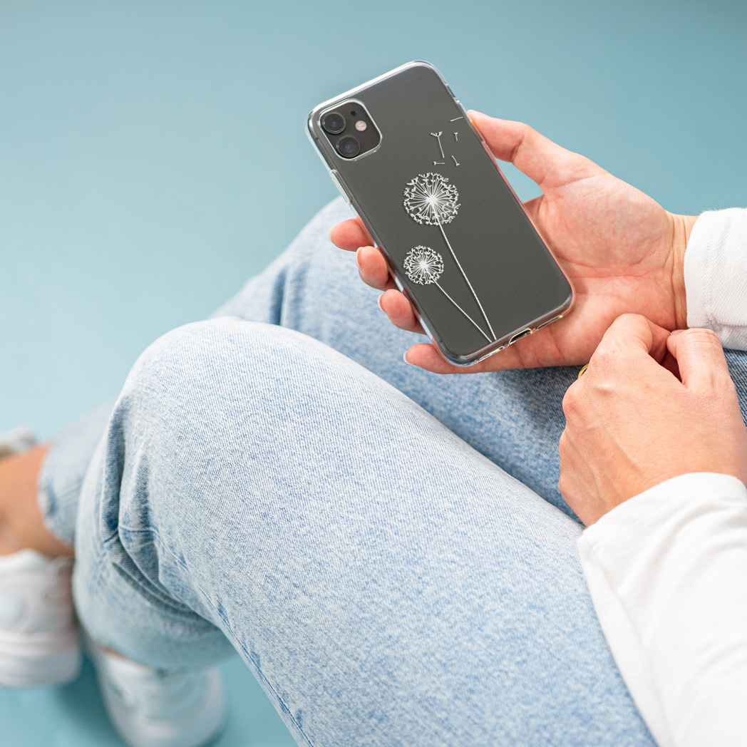 Una persona tiene in mano un telefono con una custodia a forma di dente di leone. La persona indossa jeans e una camicia bianca. Lo sfondo è azzurro.