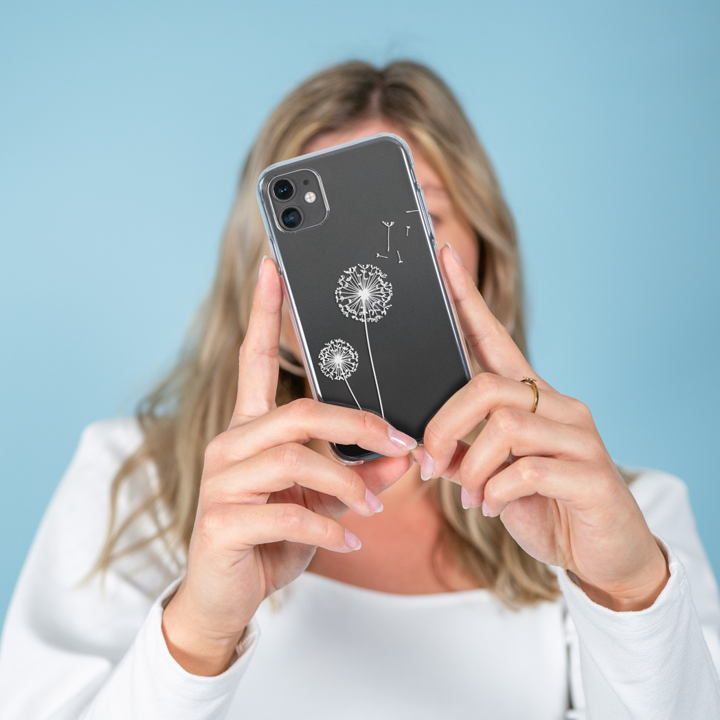 Donna che tiene in mano un telefono con un disegno di dente di leone. Lo sfondo è blu. Indossa un top bianco e un anello d'oro.