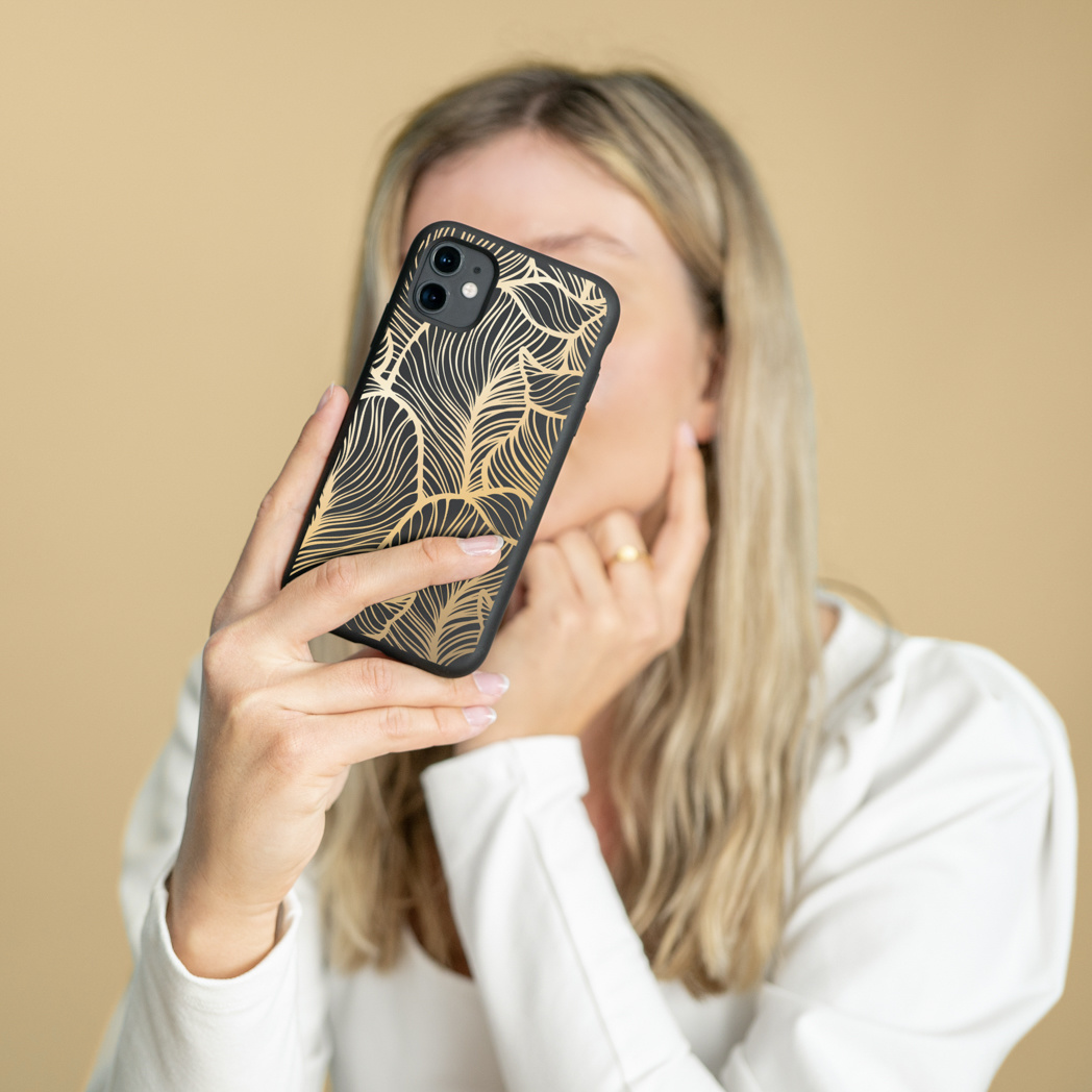 Vrouw houdt een telefoon vast met een zwarte hoes en een gouden veermotief. Ze draagt een wit overhemd en een gouden ring. Beige achtergrond.