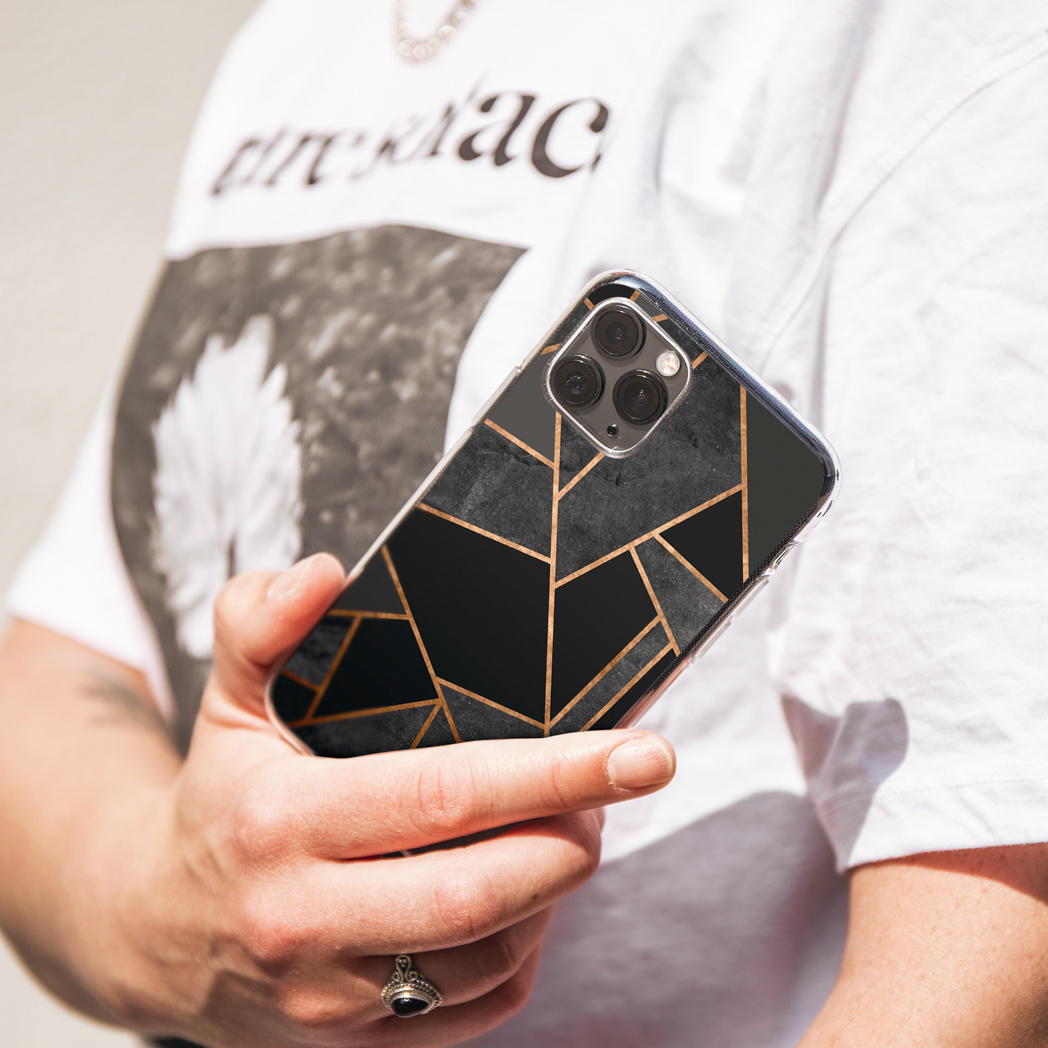 Una persona tiene in mano un telefono con una custodia geometrica nera e oro. La persona indossa una camicia bianca e un anello d'argento.