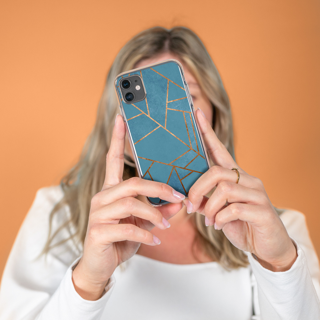 Donna che tiene in mano un telefono con una custodia blu dal design geometrico. Lo sfondo è arancione. Indossa una camicia bianca e un anello d'oro.