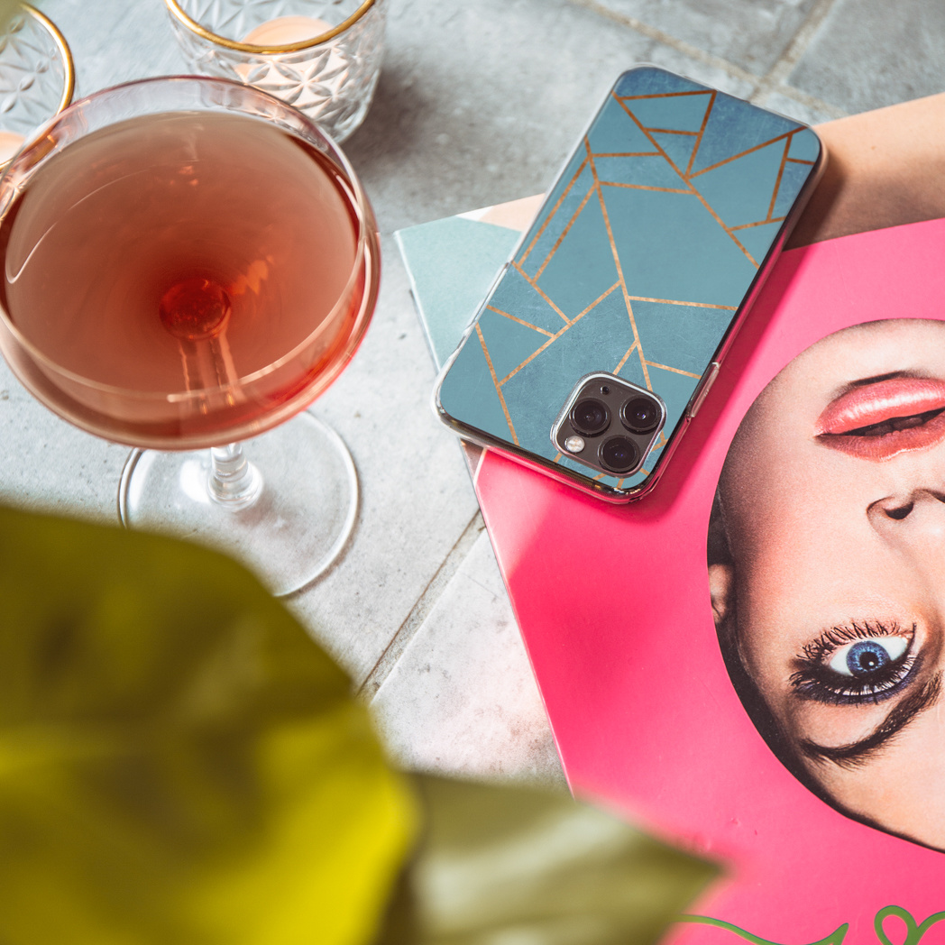Un bicchiere da cocktail con una bevanda rosa, un telefono con una custodia blu e oro e una rivista con il volto di una donna.