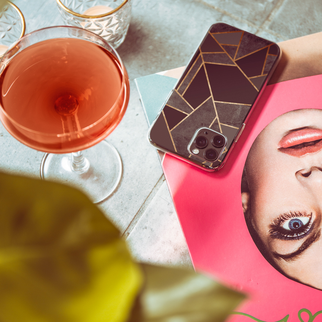 Un cocktail, una custodia per telefono e una rivista sono su un tavolo. La custodia ha un design geometrico. La rivista mostra un volto.