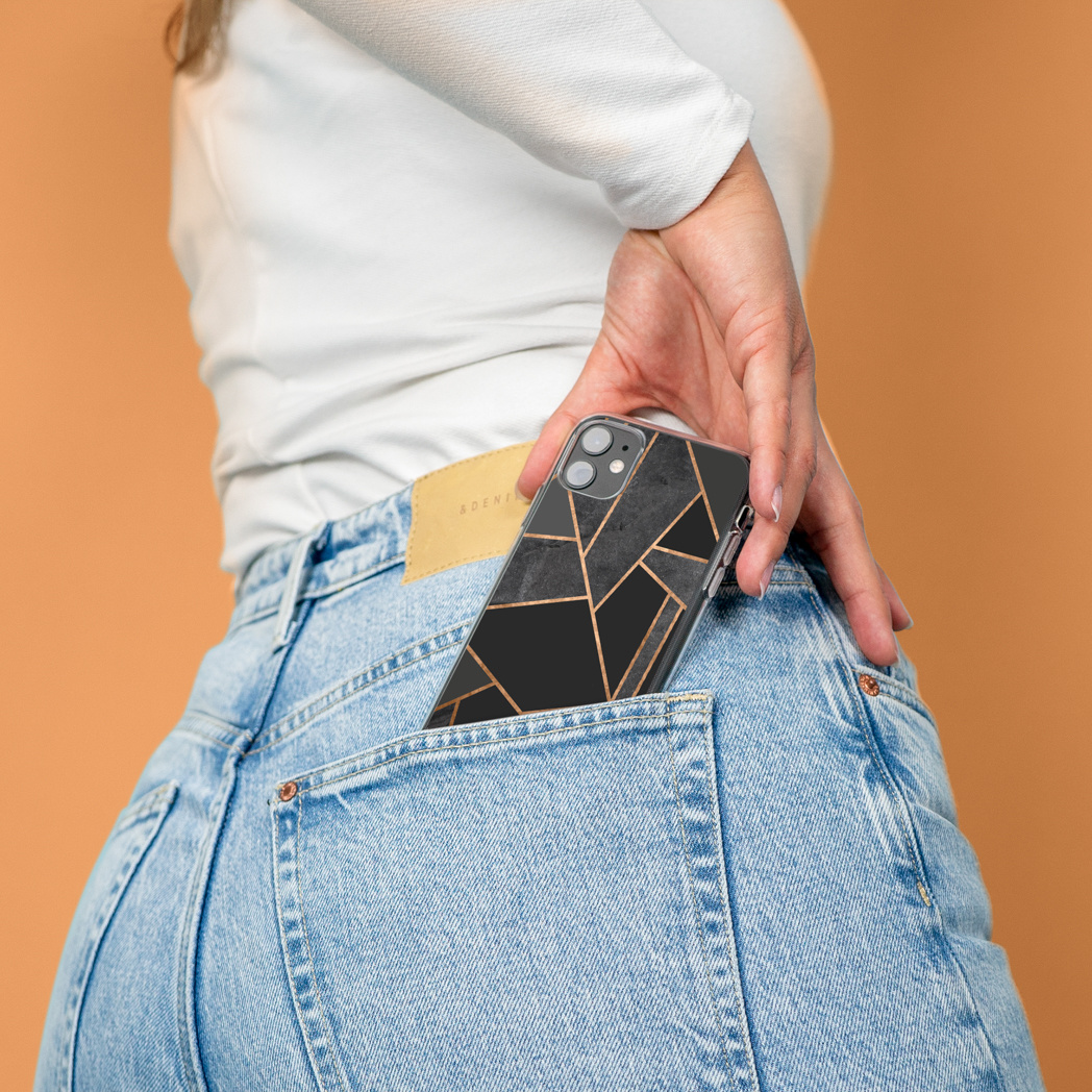 Una mano tiene un telefono con un design geometrico nero e oro, parzialmente in una tasca di jeans.