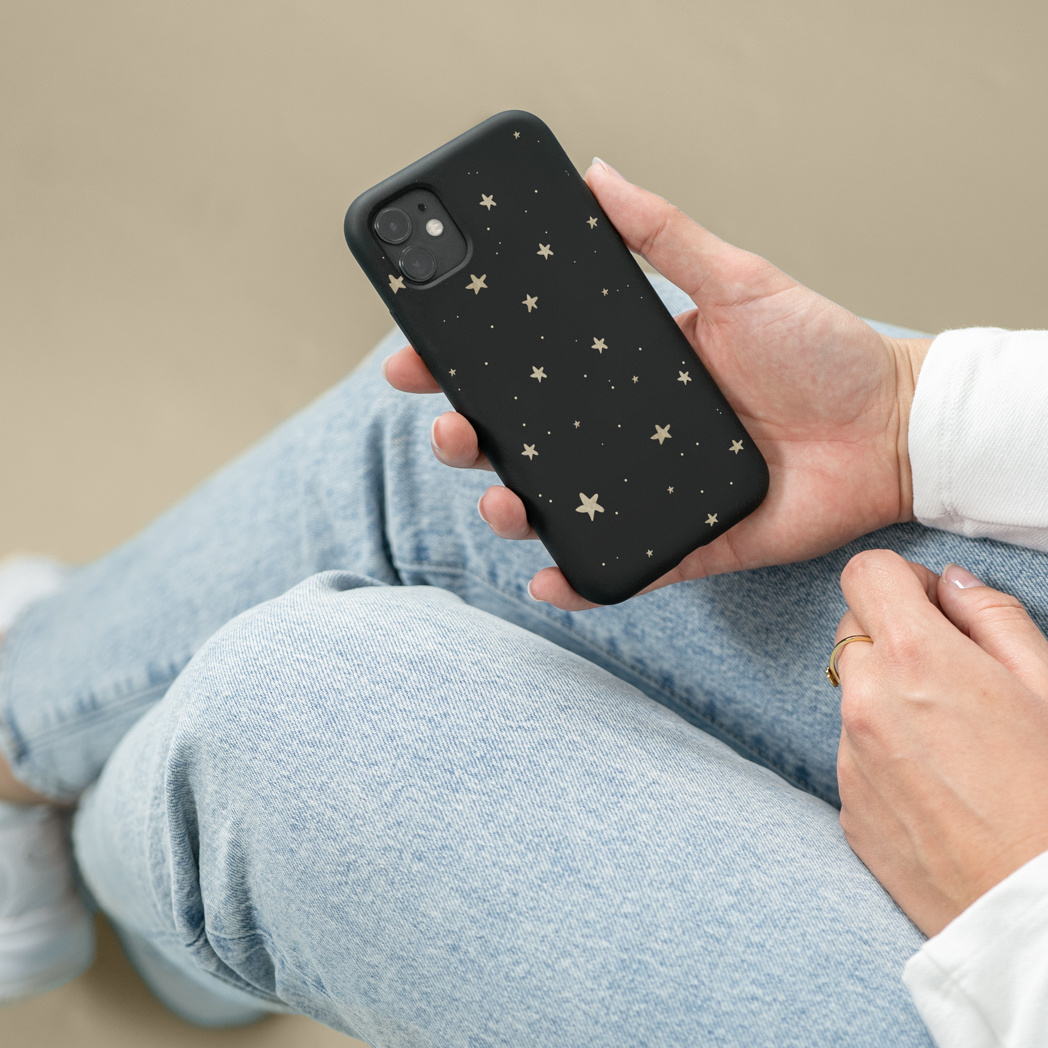 Una persona tiene in mano un telefono nero con stelle dorate. La persona indossa jeans azzurri, una camicia bianca e un anello d'oro.