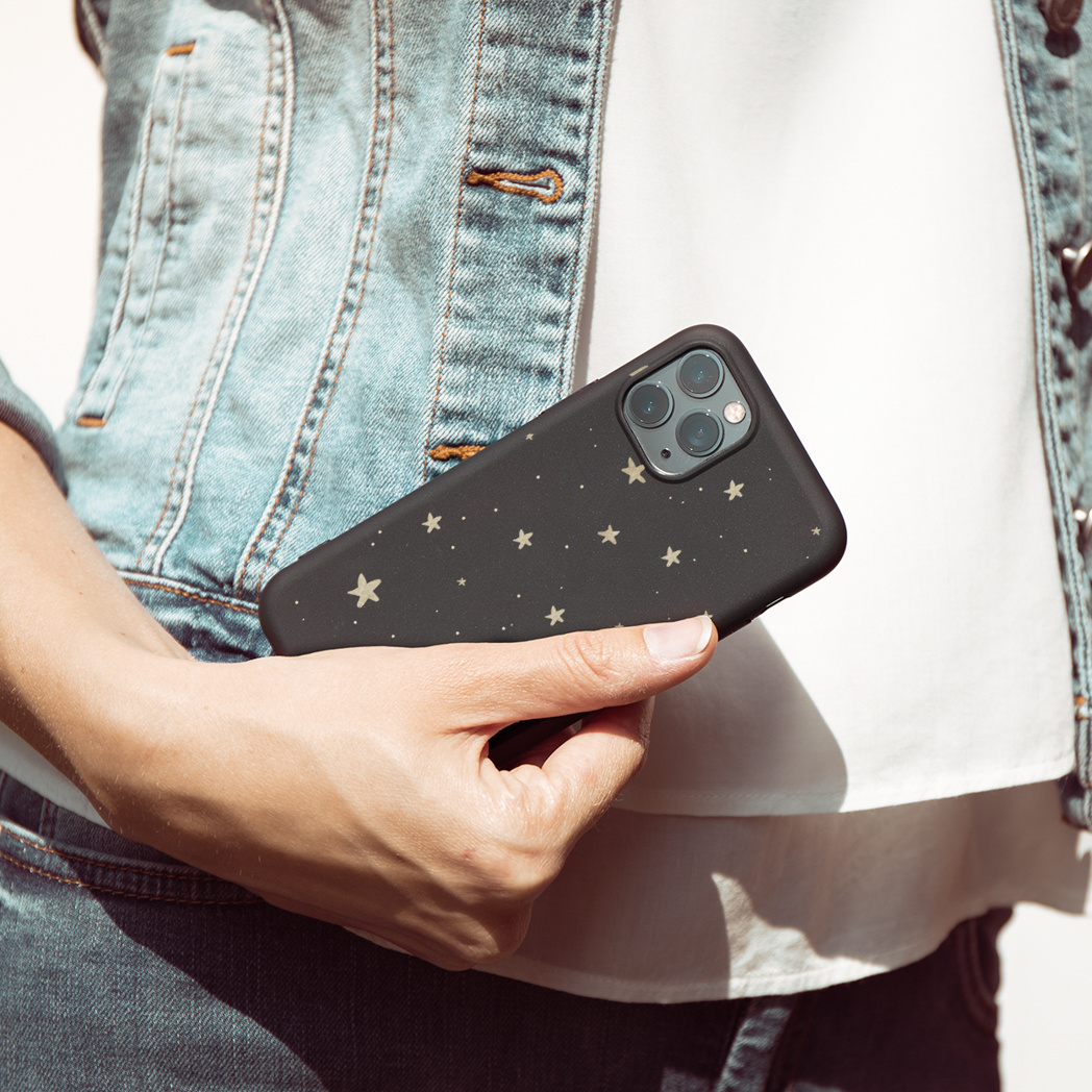 Una persona tiene in mano un telefono. La custodia del telefono è nera con stelle dorate. La persona indossa una giacca di jeans e una camicia bianca.
