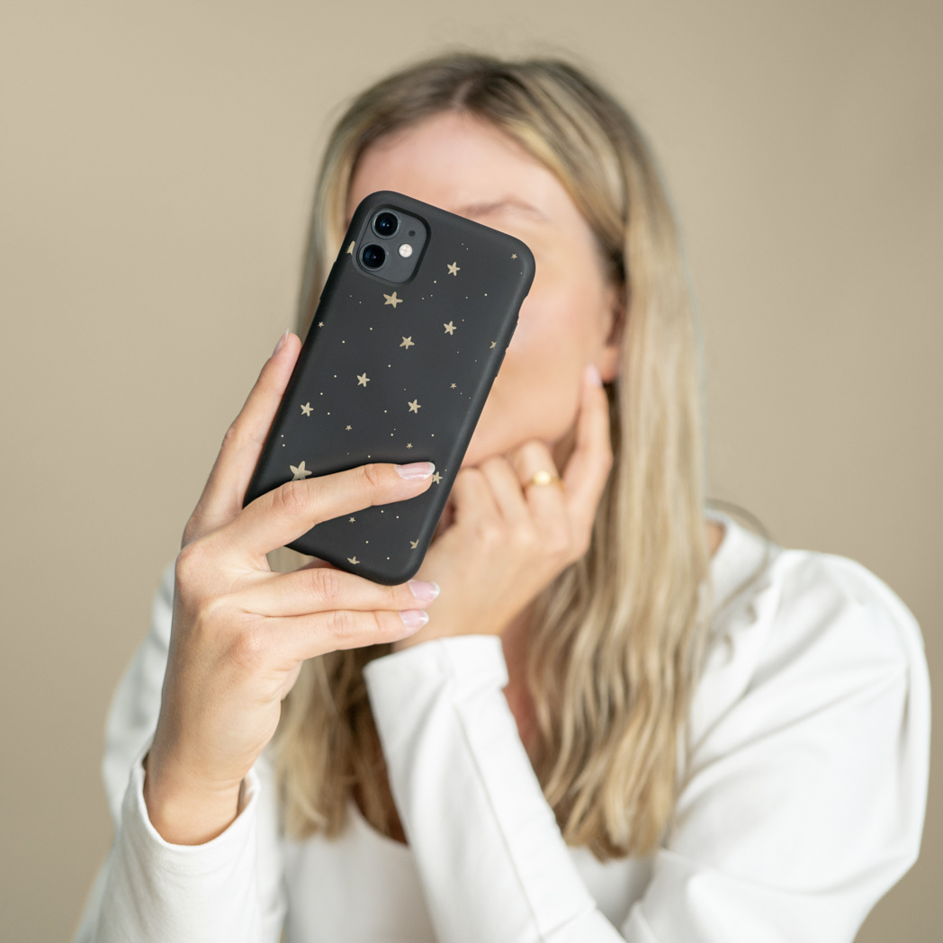 Una donna tiene in mano un telefono nero con stelle dorate. Indossa una camicia bianca e un anello d'oro. Il suo viso è parzialmente visibile.