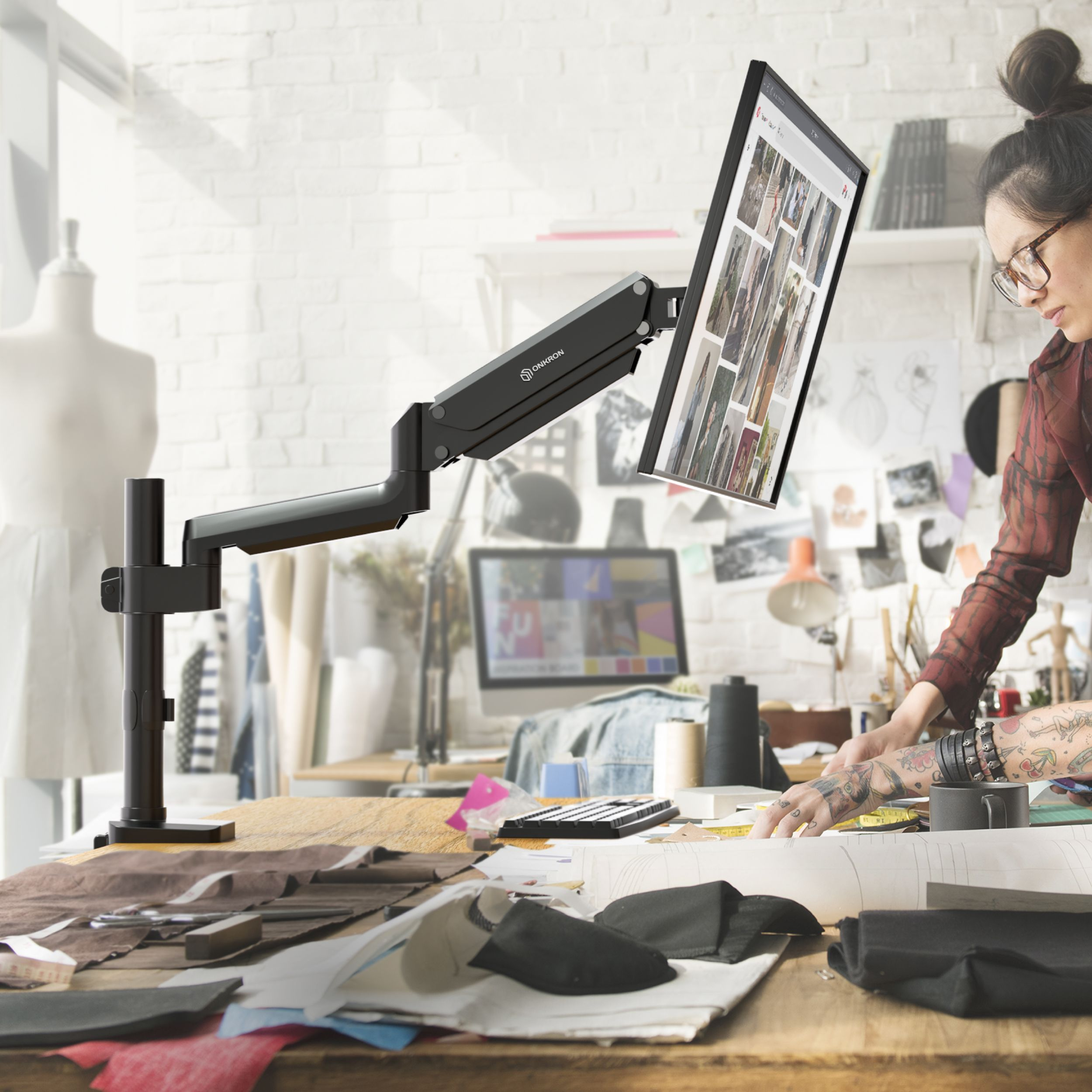 Kobieta w okularach pracuje przy biurku z komputerem. Ramię monitora trzyma ekran. Na biurku jest materiał i narzędzia.