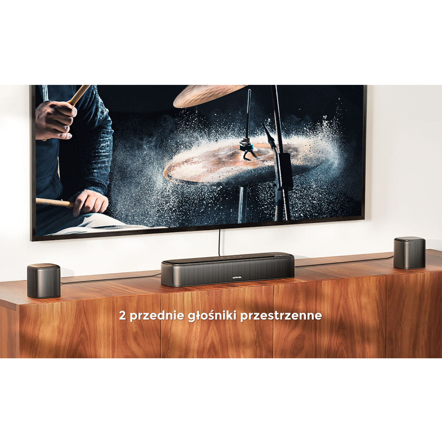 Ekran telewizora pokazuje perkusistę. Poniżej soundbar z dwoma głośnikami. Drewniana szafka trzyma zestaw.