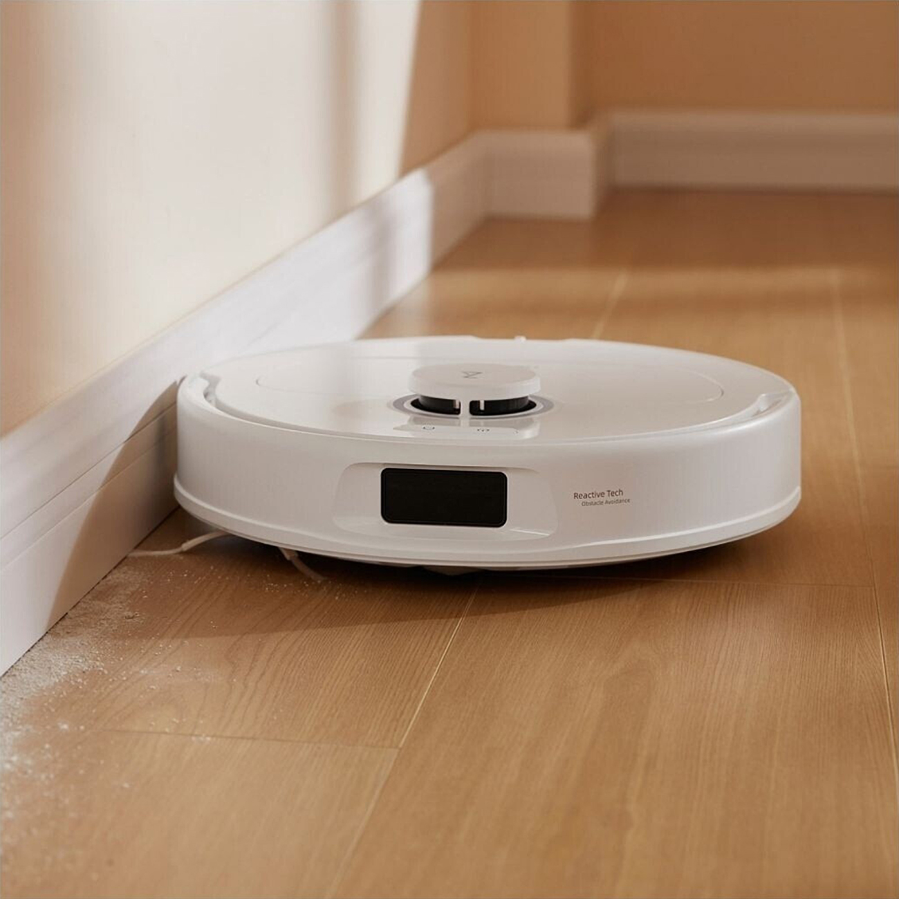 Un aspirapolvere robot bianco su pavimenti in legno, vicino a un muro. Ha uno schermo e un pulsante. Un po' di polvere è visibile.
