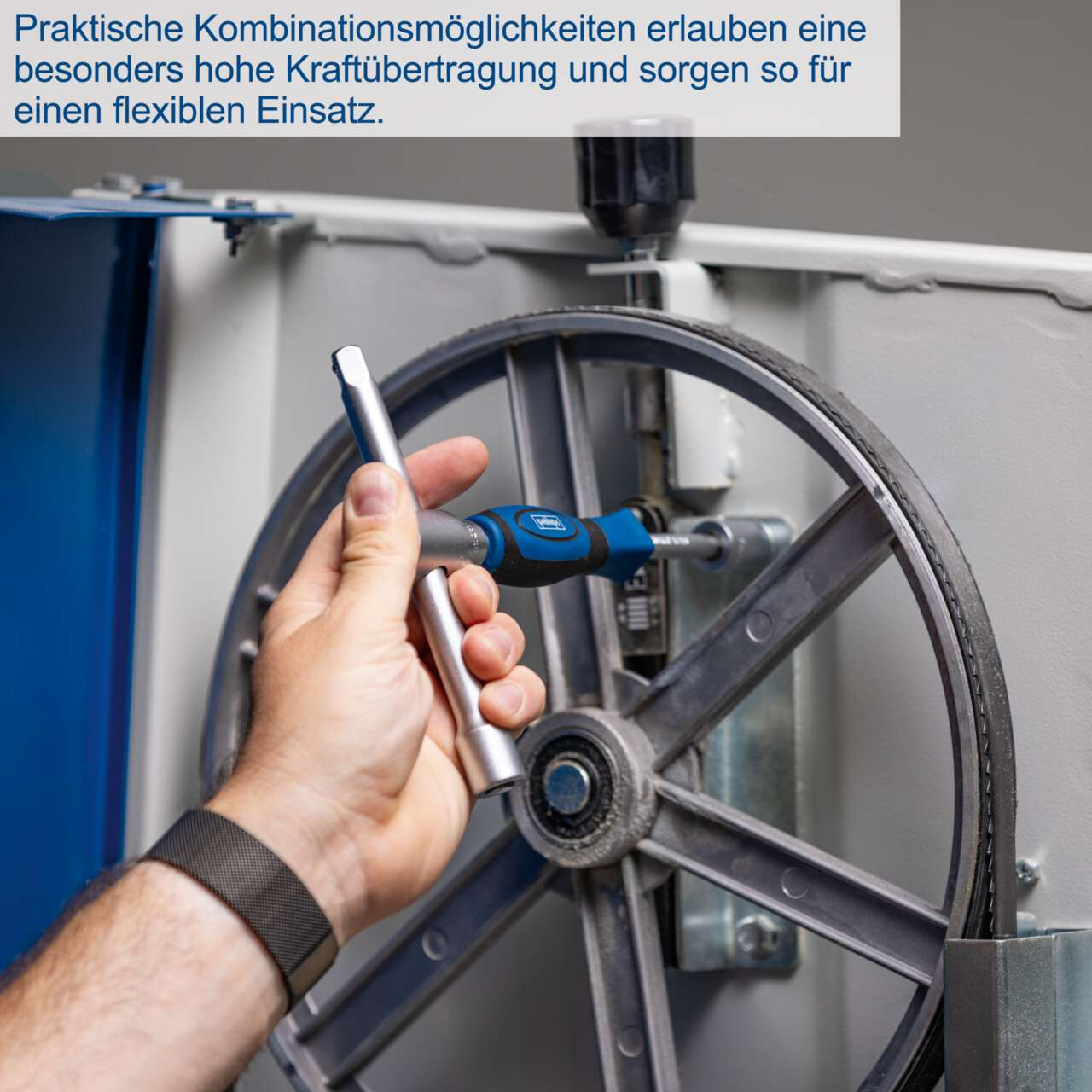 A hand holding a blue and silver tool is tightening a bolt on a large wheel. The background is a machine with a belt.