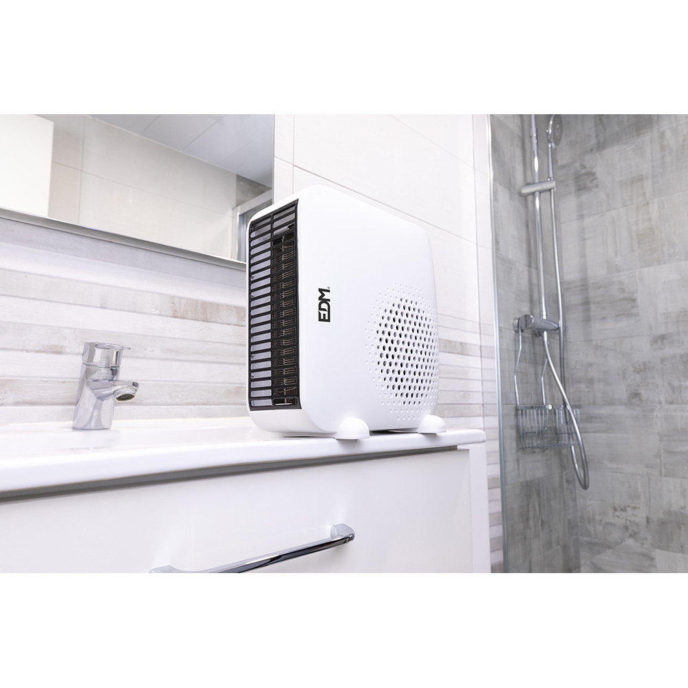 Un calentador eléctrico blanco con rejillas negras está sobre un lavabo blanco. Un grifo plateado, espejo y ducha están en el fondo.