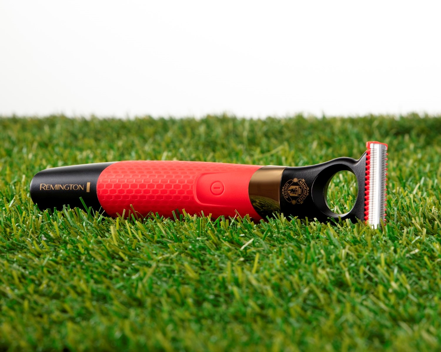 Una afeitadora Remington roja y negra está sobre césped artificial verde. La afeitadora tiene una cuchilla metálica y un logo del Manchester United.