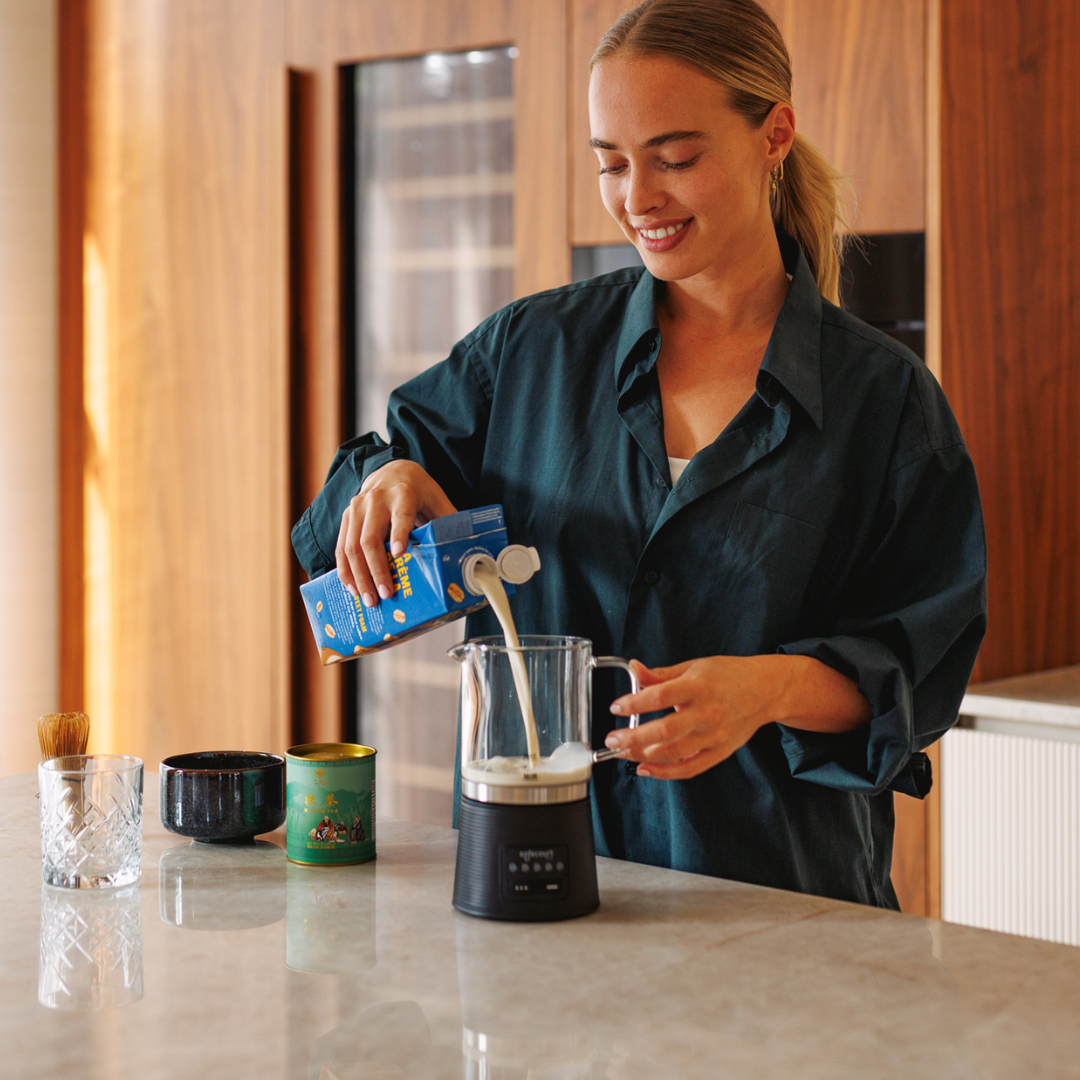 Een vrouw giet melk uit een blauwe doos in een glazen opschuimer. Andere items zijn kommen, een glas en een borstel.