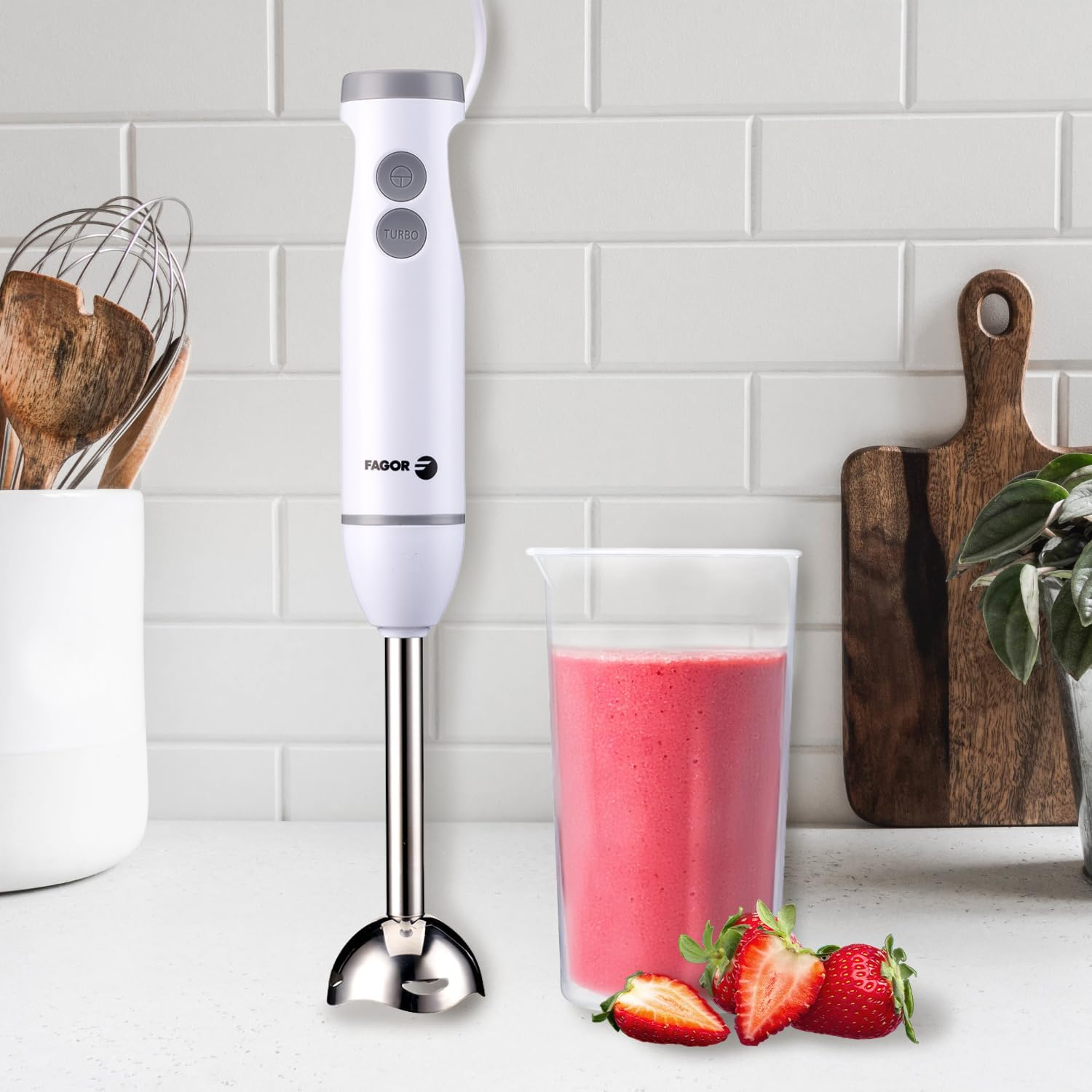 Una licuadora blanca con un vaso de batido rosa y fresas, sobre una encimera blanca. Fondo: pared de ladrillo blanco y tabla de madera.