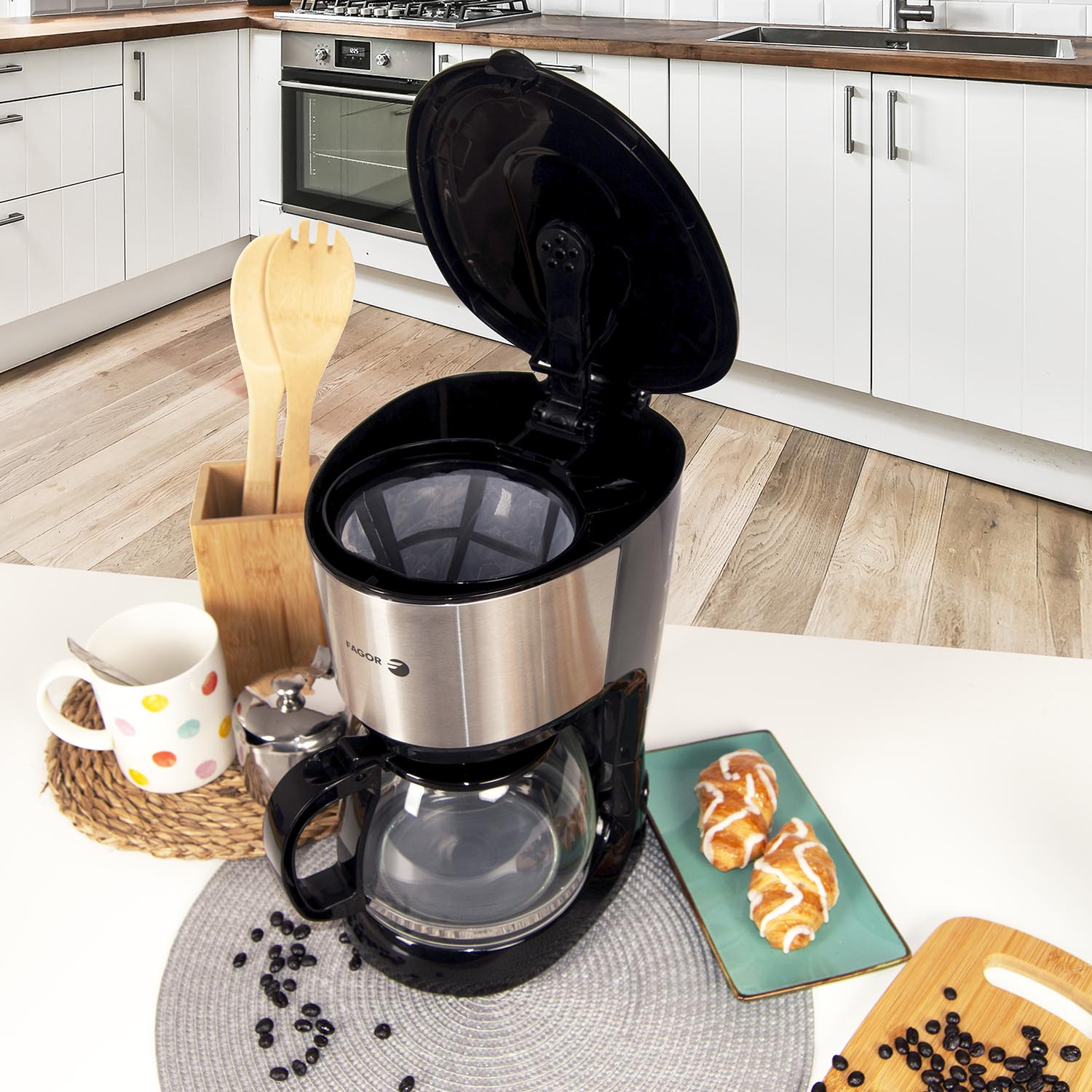 Cafetera sobre una mesa con pasteles, taza y granos. Cocina al fondo.