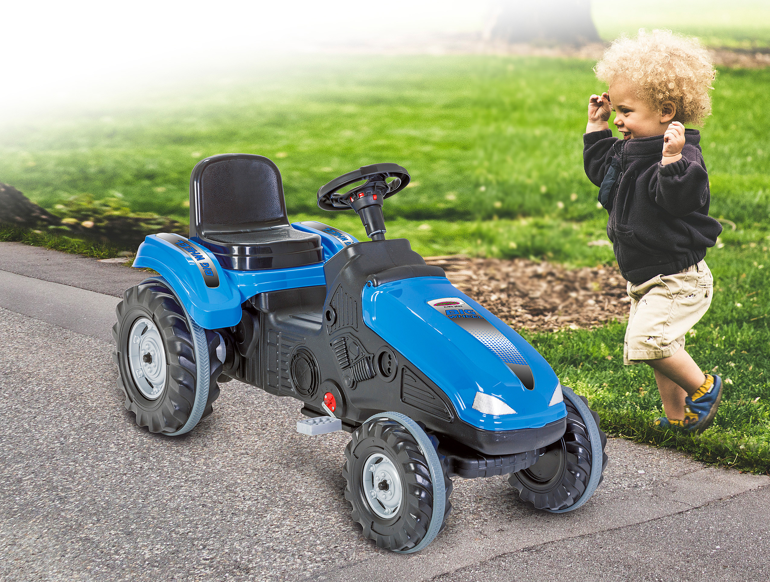 Blauer Spielzeugtraktor auf einem gepflasterten Weg. Ein Kind mit lockigem Haar steht im Gras und lächelt.