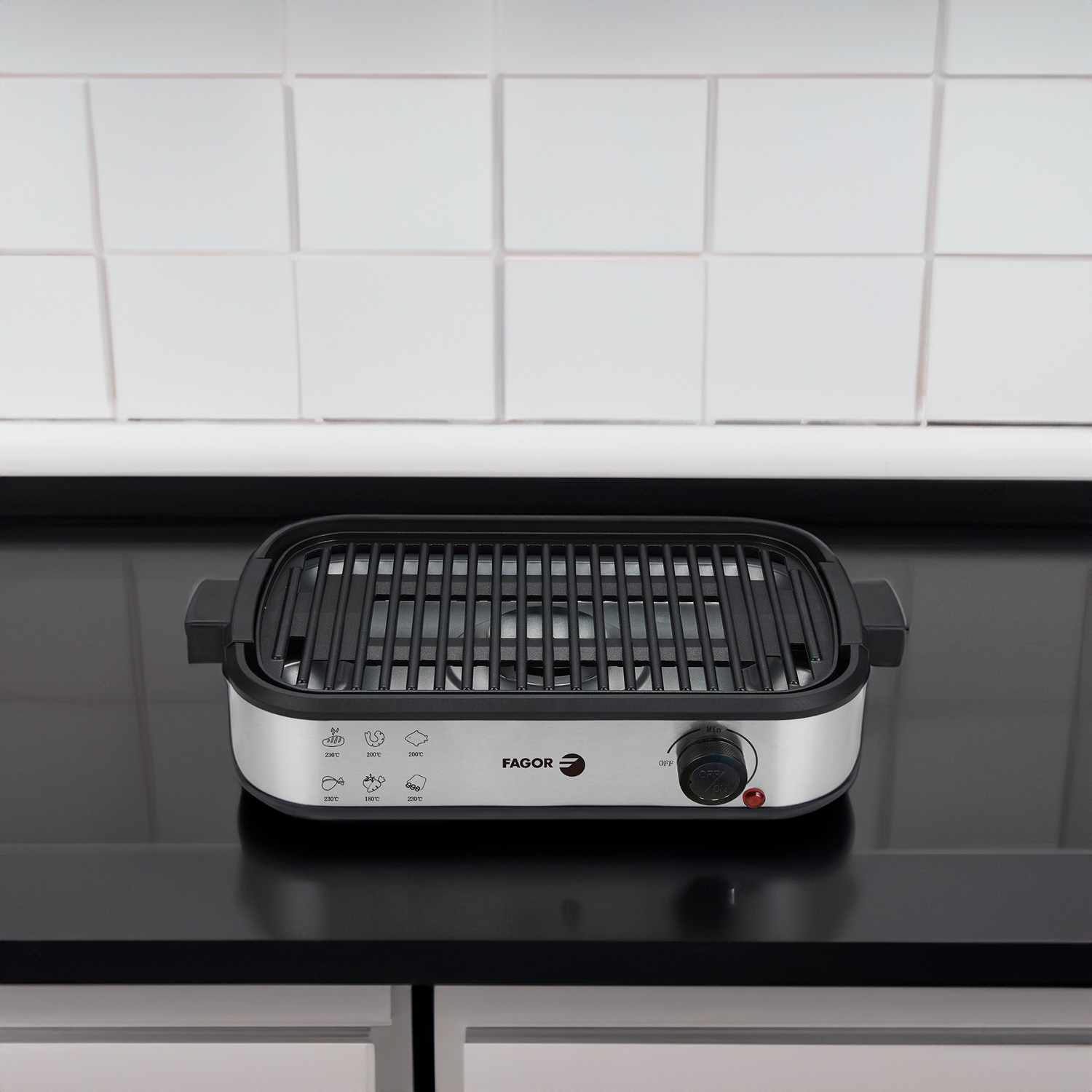Una parrilla eléctrica Fagor con parte superior negra, base plateada y perilla de control de temperatura, sobre una superficie negra y fondo de azulejos blancos.