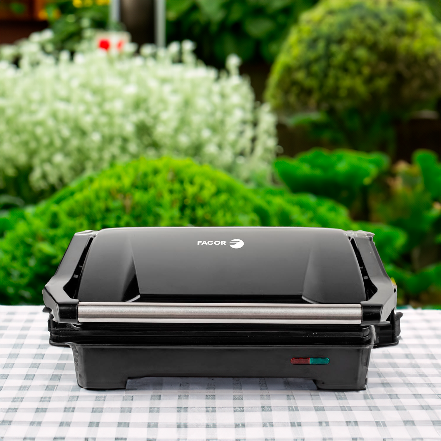 Una parrilla Fagor negra está sobre una mesa a cuadros. Hay follaje verde y blanco en el fondo. La parrilla está cerrada.