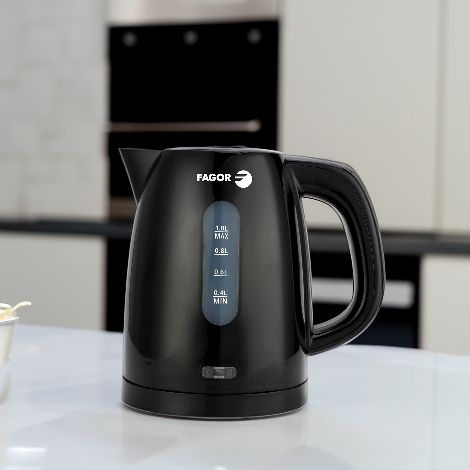 Un bollitore elettrico nero Fagor su una superficie bianca. Ha un manico, un indicatore del livello dell'acqua ed è in un ambiente cucina.
