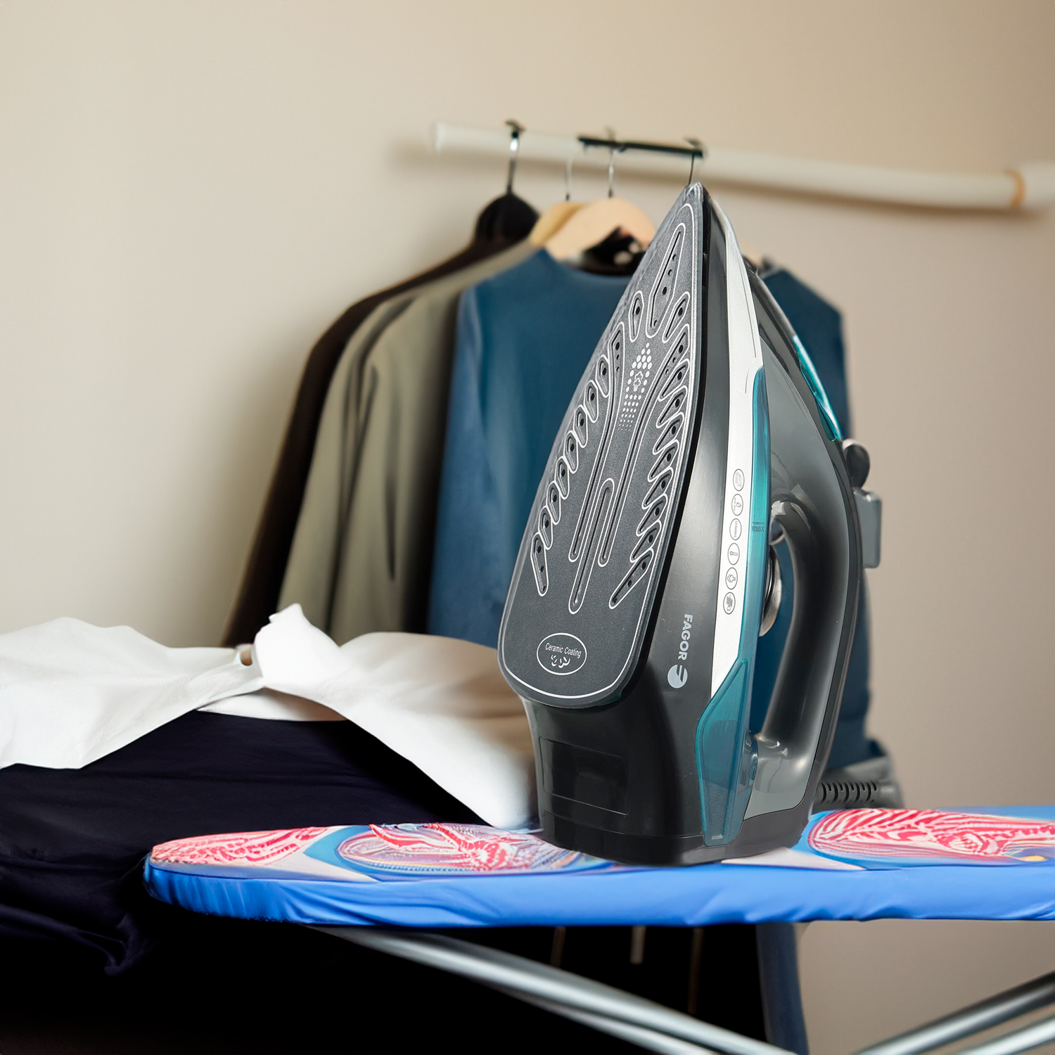 Una plancha está sobre una tabla de planchar azul con ropa. En el fondo cuelgan camisas y chaquetas.