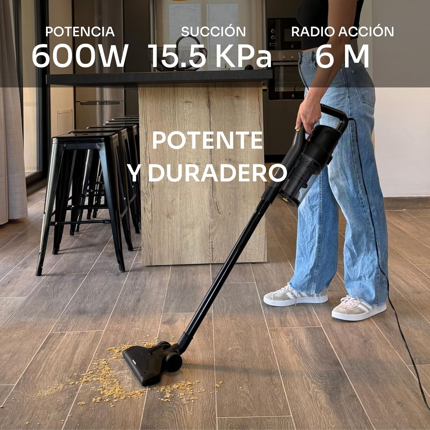 Una persona aspira un piso de madera con migas. La aspiradora es negra. La persona usa jeans y zapatillas blancas.