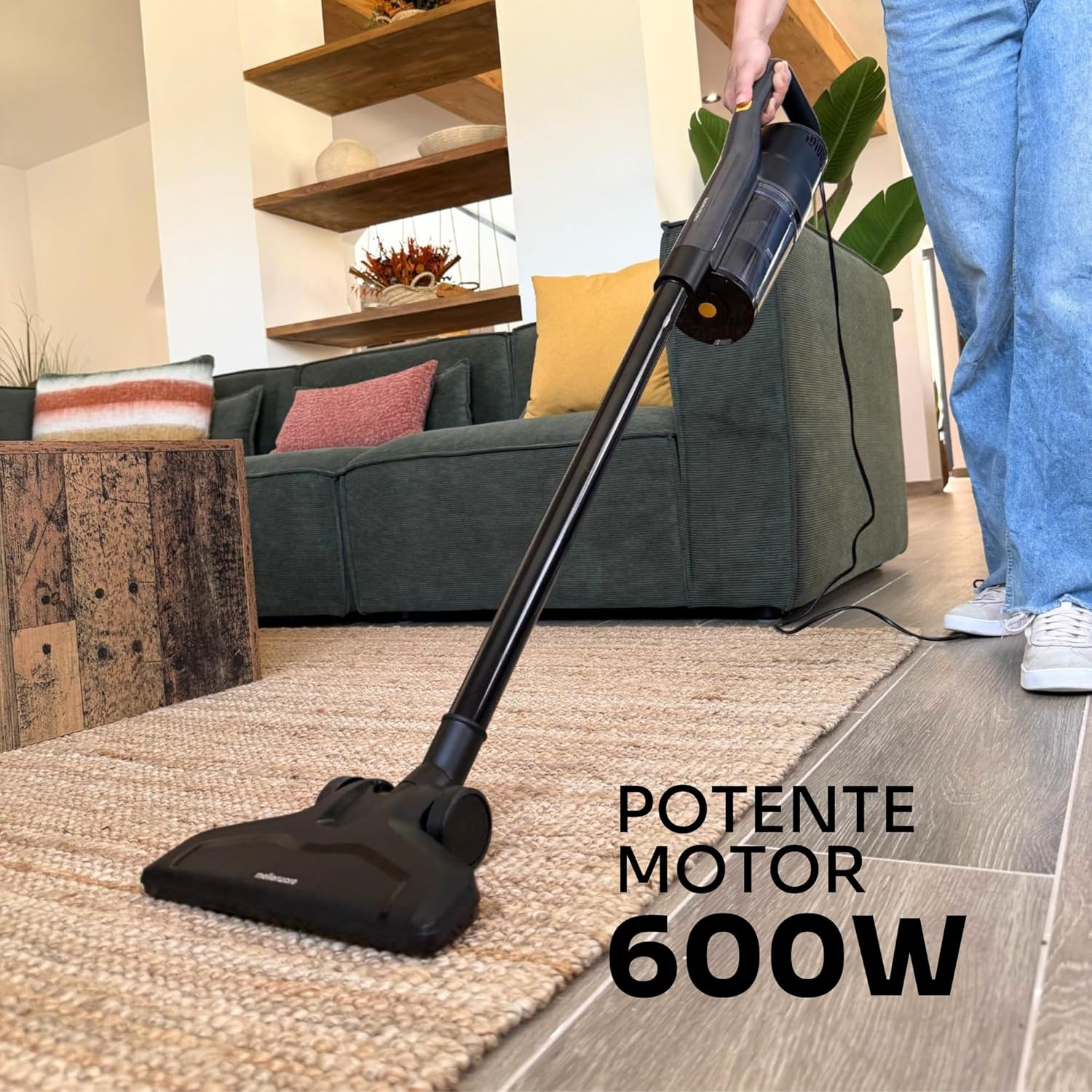 Una persona usa una aspiradora negra en una alfombra. En el fondo, un sofá, estanterías y el texto 'POTENTE MOTOR 600W'.