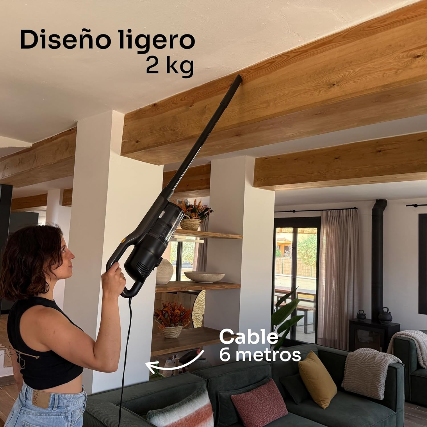 Mujer usando una aspiradora negra en una viga de madera. La escena incluye un sofá verde, estantes y una puerta abierta.