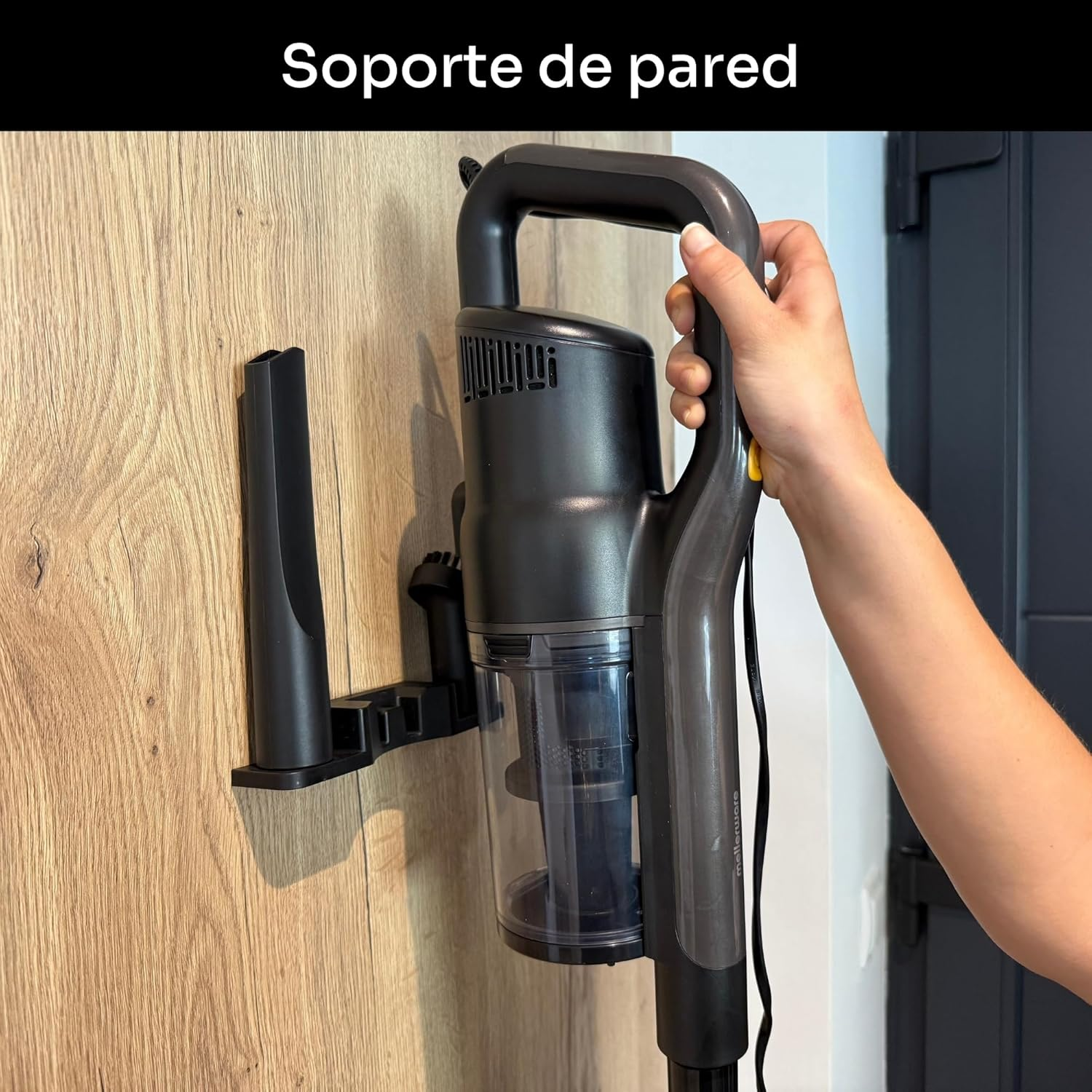 Una aspiradora negra sostenida por una persona. Está montada en una pared de madera. El fondo es una pared blanca y una puerta oscura.