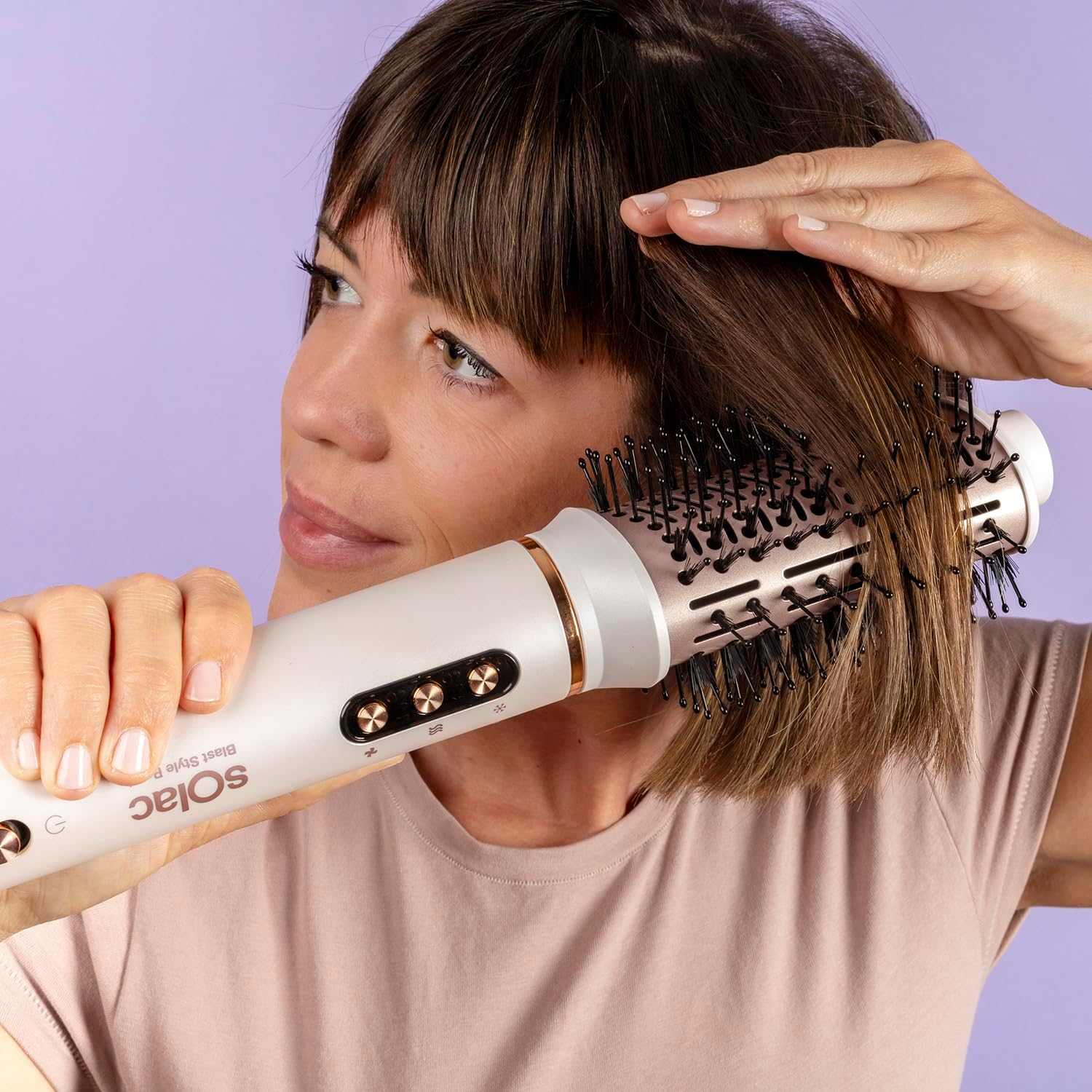 Una mujer peina su cabello castaño con un cepillo blanco y dorado. Lleva una camisa rosa. El fondo es morado.