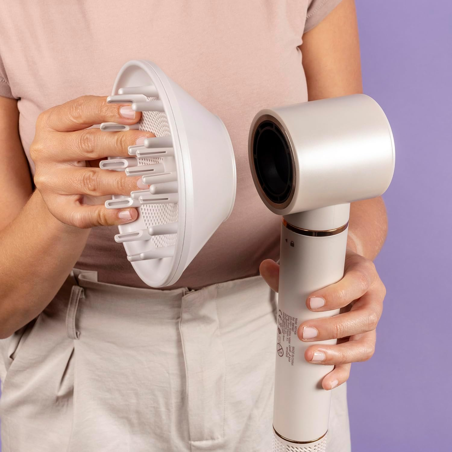 Una persona sostiene un secador de pelo blanco con un accesorio difusor. La persona lleva una camiseta y pantalones claros, con fondo morado.