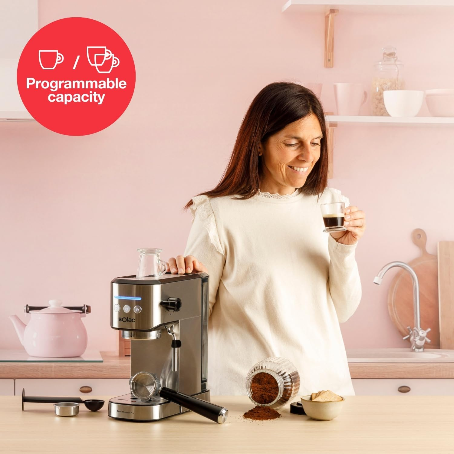Una mujer en una cocina sosteniendo café, una cafetera y café molido en una mesa. Paredes y objetos rosas en el fondo.