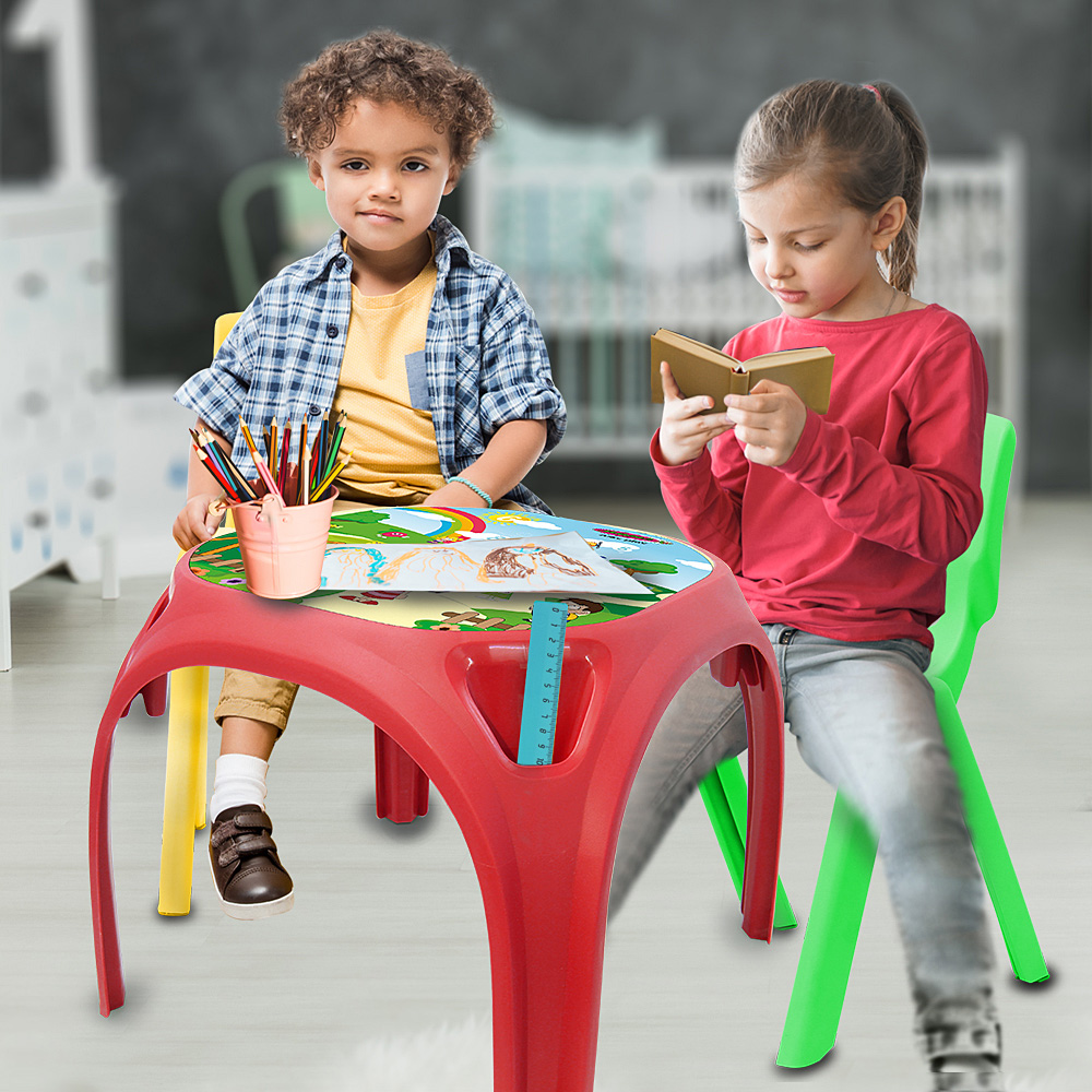 Zwei Kinder sitzen an einem roten Tisch. Eines liest ein Buch, das andere schaut in die Kamera. Farbstifte in einem Becher sind sichtbar.