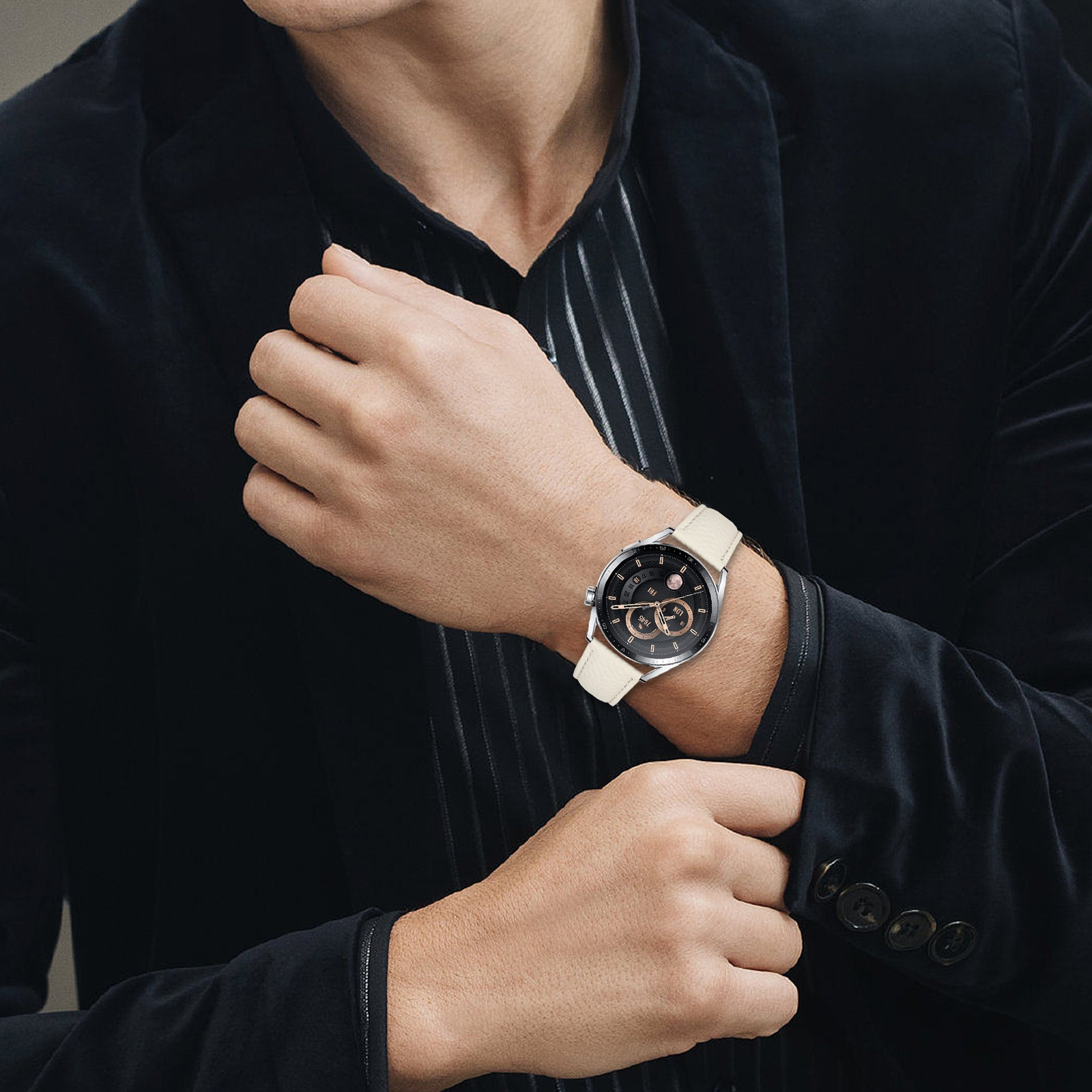 Un uomo in abito nero sistema la manica, rivelando un orologio da polso con cinturino color crema e quadrante nero con dettagli dorati.